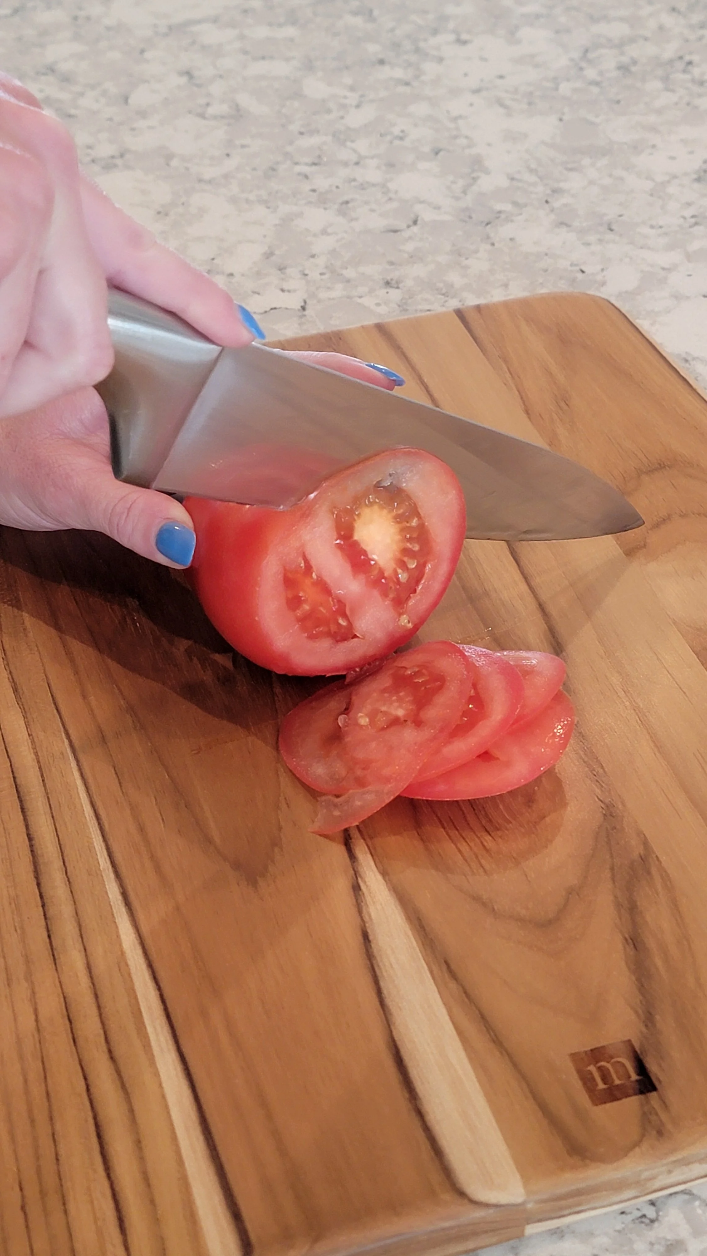 Person slicing a tomato on a wooden cutting board with a chef's knife.