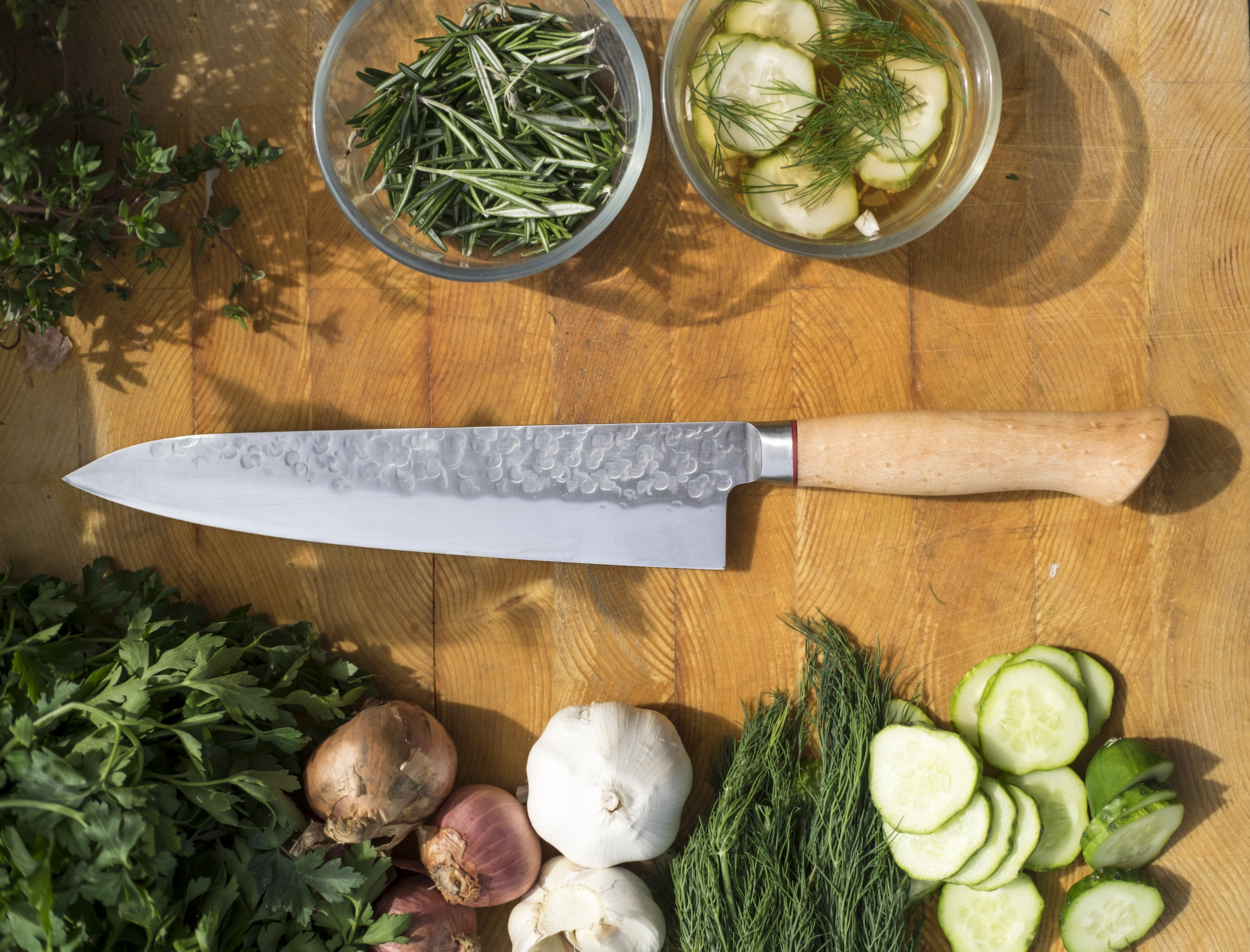 Fresh herbs and vegetables arranged on a wooden surface, including sliced cucumbers, garlic, shallots, dill, parsley, and a knife with a patterned blade and light-colored handle.