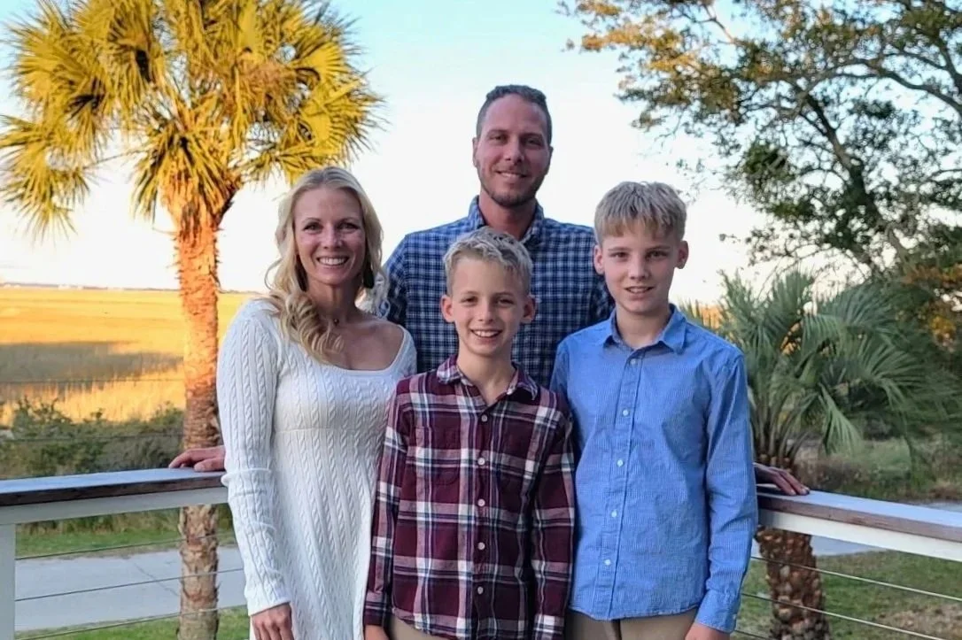 A family of five standing outdoors on a balcony with palm trees and a field in the background. The family includes a woman, a man, and three boys, all smiling at the camera.