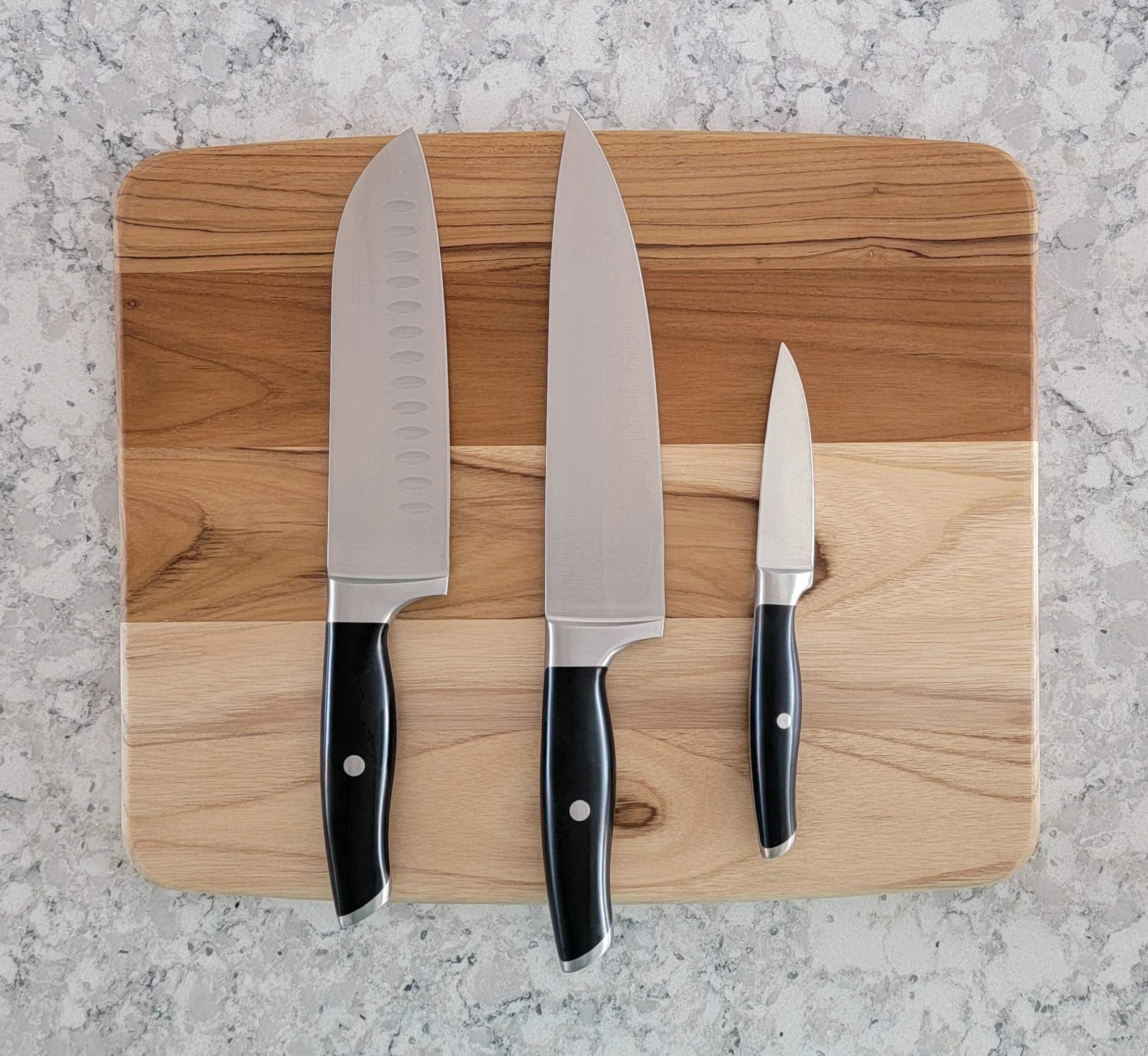 Three stainless steel knives with black handles on a wooden cutting board, placed on a gray stone countertop.