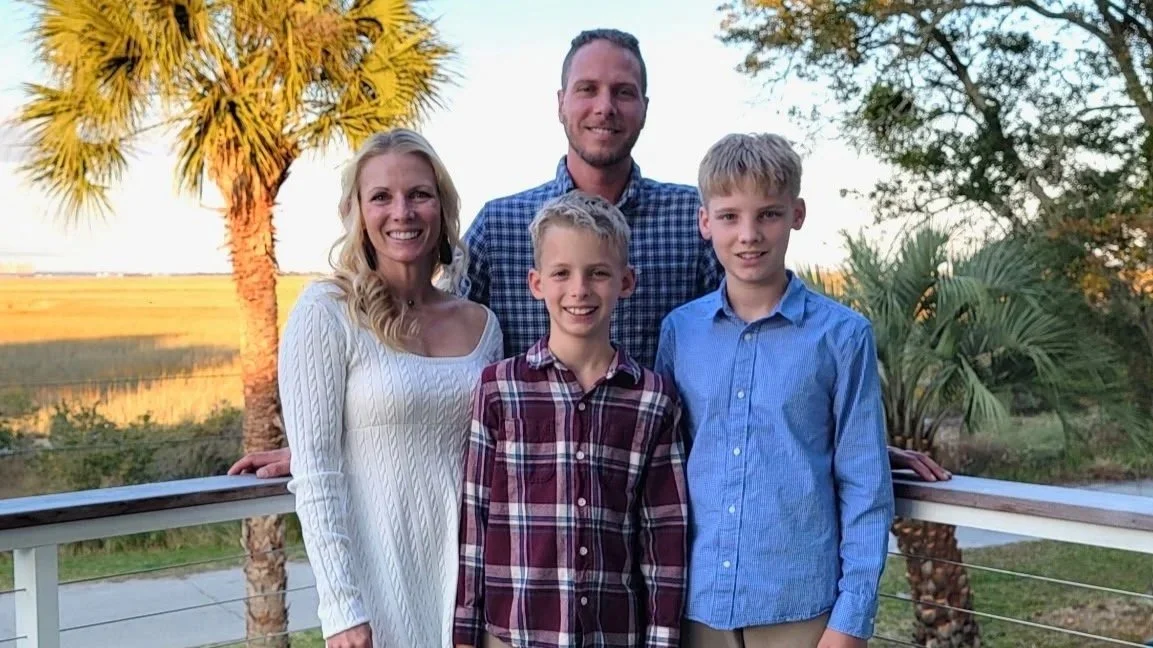 Family of four smiling outdoors on a balcony with palm trees and open field in the background.