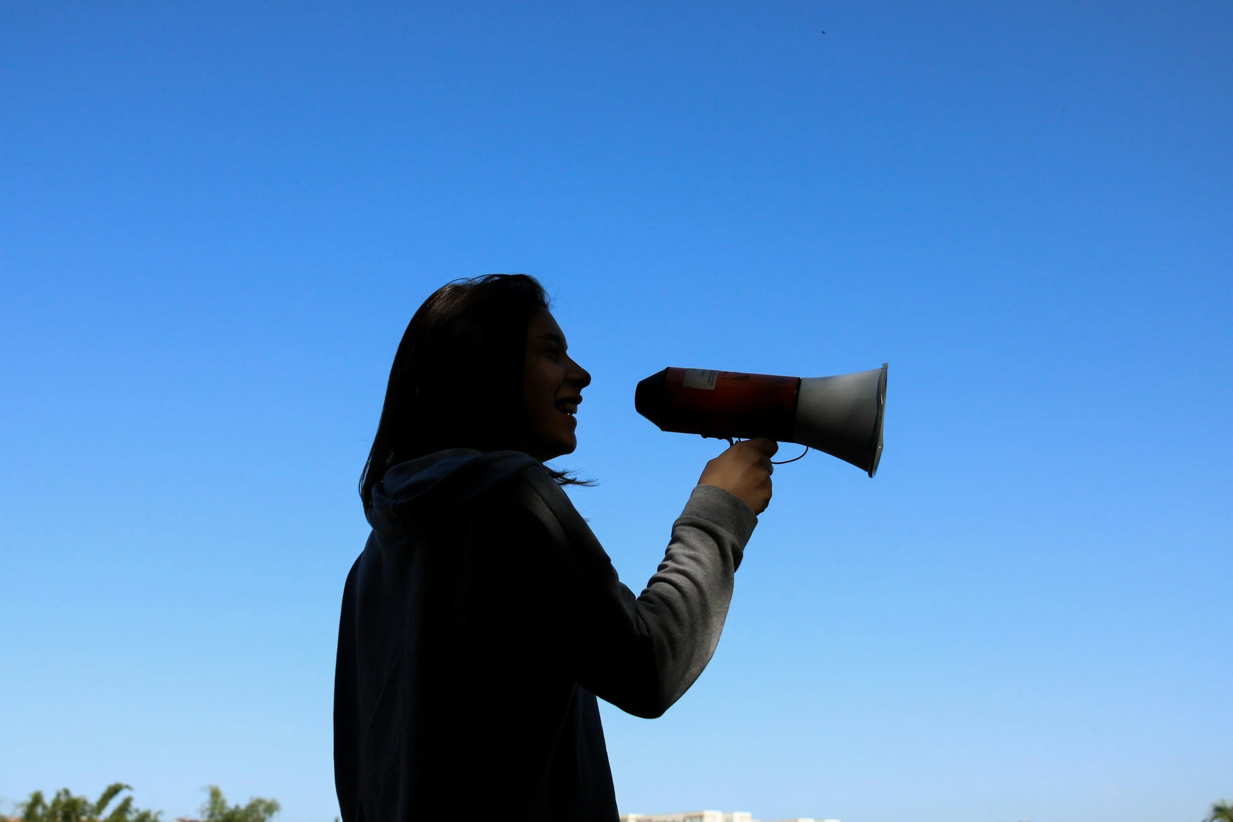 A person holding a megaphone and smiling, silhouette against a clear blue sky.