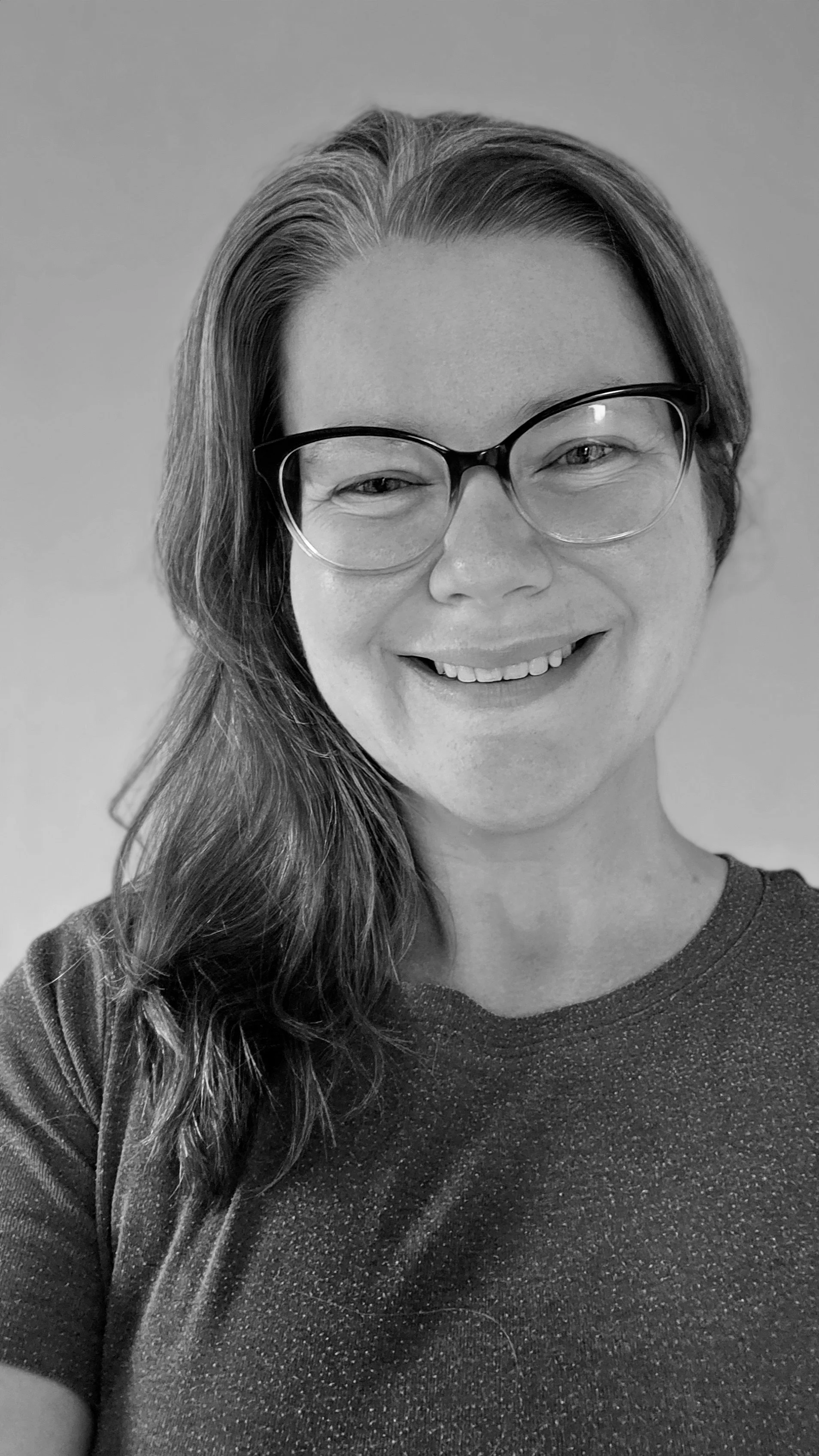 Black and white photo of Amy Dove, a woman with glasses smiling, wearing a dark shirt, with long wavy hair over her shoulder.