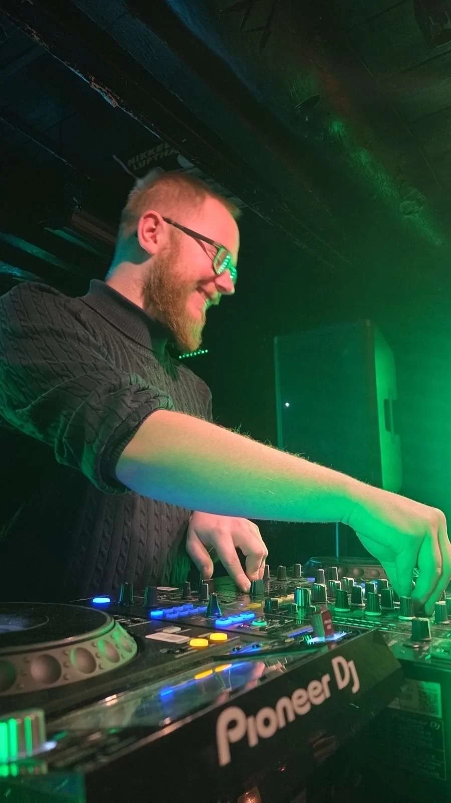 A DJ with glasses, a beard, and a black sweater is smiling while operating a DJ controller in a dimly lit club with green lighting.