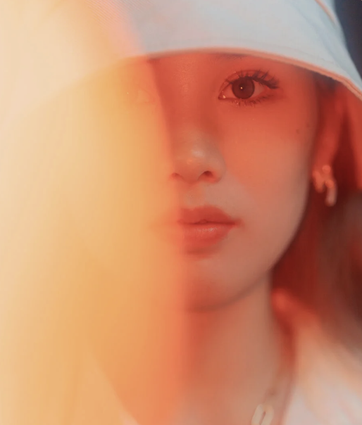 Close-up of a young woman with fair skin, one eye visible, wearing a light-colored bucket hat and earrings, with warm reddish lighting.