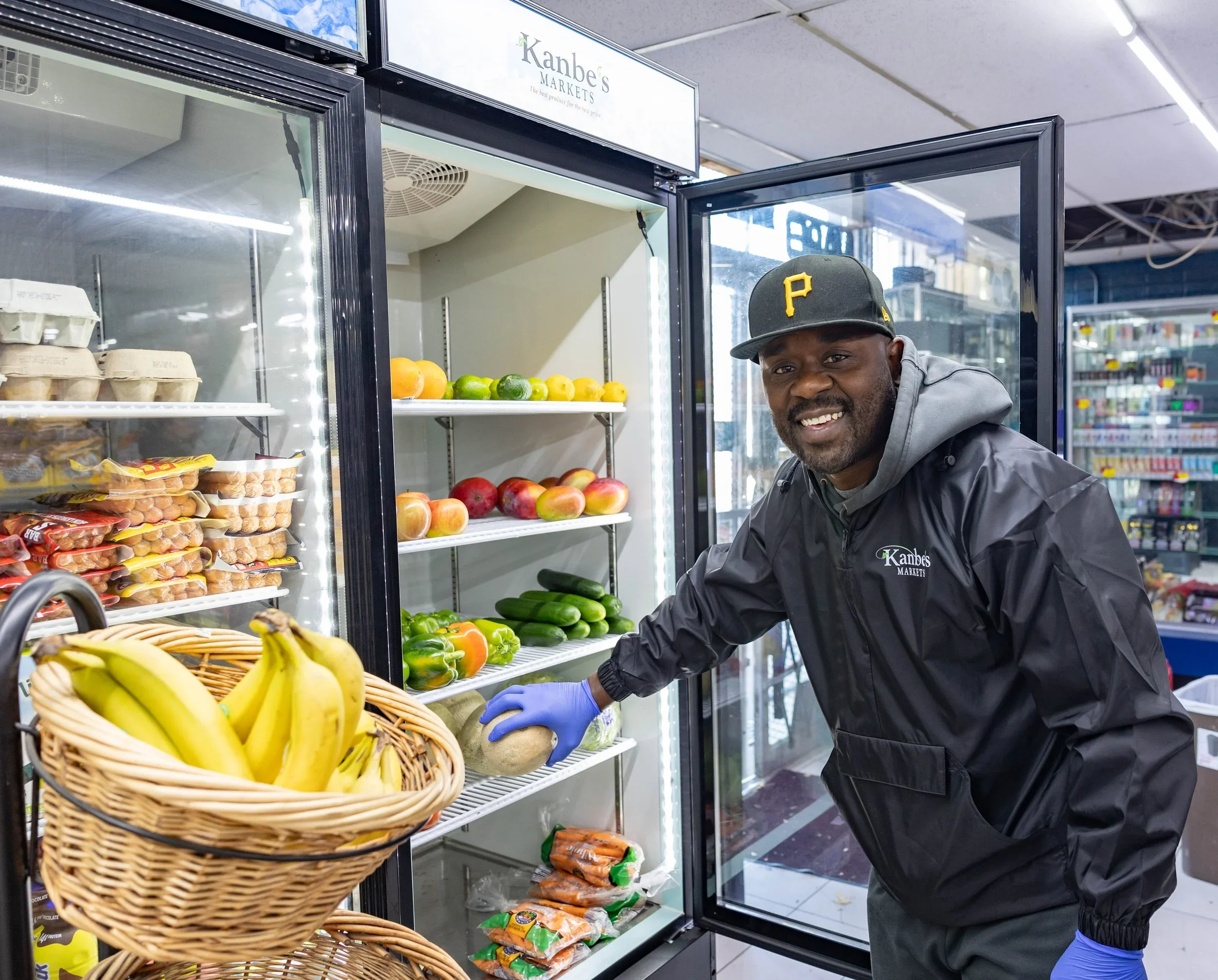 A man wearing a black jacket, gray hoodie, black cap with a yellow 'P', and blue gloves shopping for fruit at a grocery store. He is placing an eggplant into a basket near a refrigerator with various fruits and vegetables.