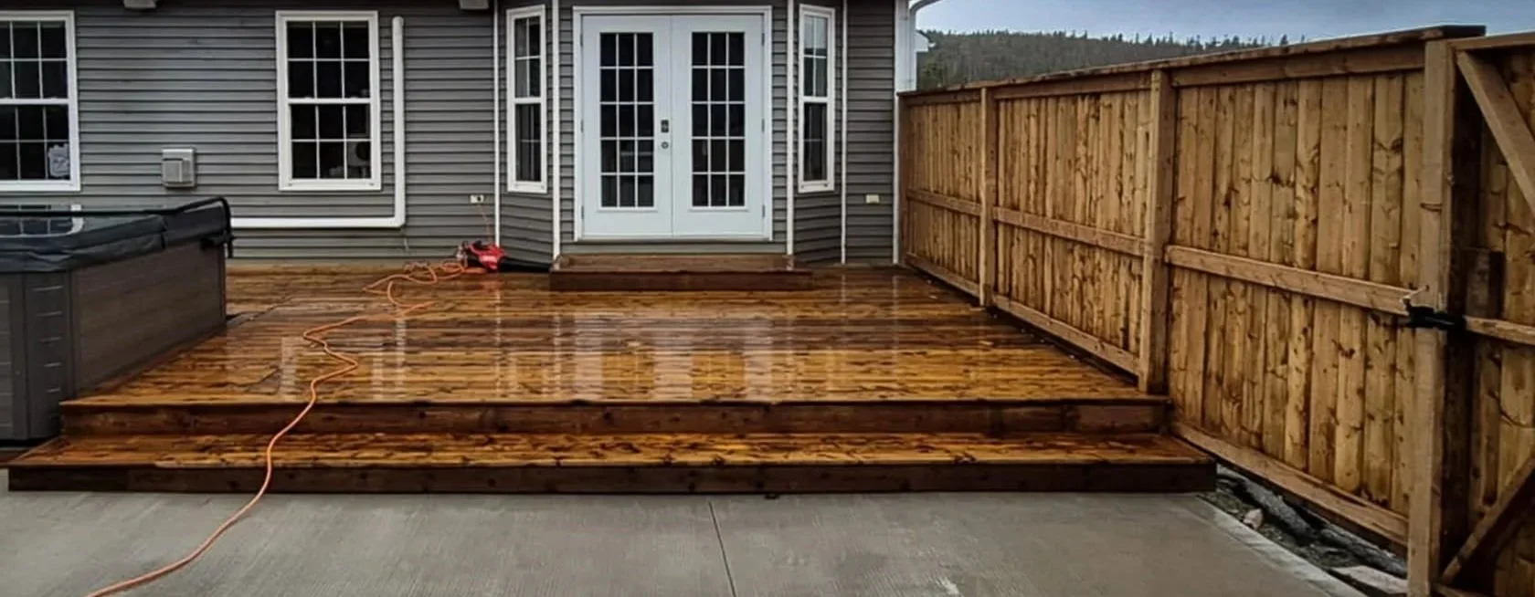 Backyard wooden deck with a railing on the right side, stairs leading down to a concrete patio, a hot tub on the left side, and a house with glass double doors and windows in the background. The deck appears wet or recently cleaned.