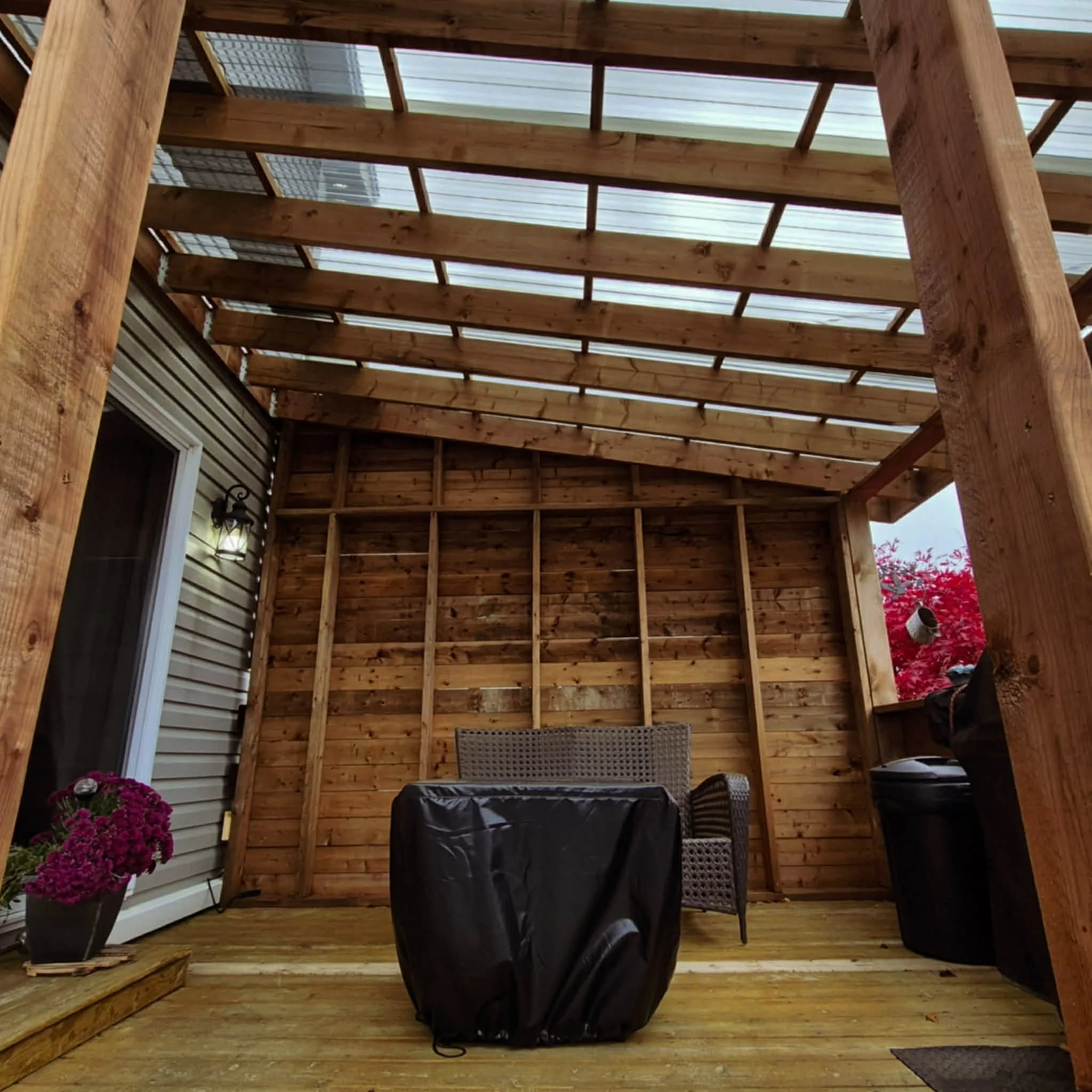 A partially enclosed outdoor wooden patio with a sloped roof, wicker furniture, potted flowers, and storage bins.