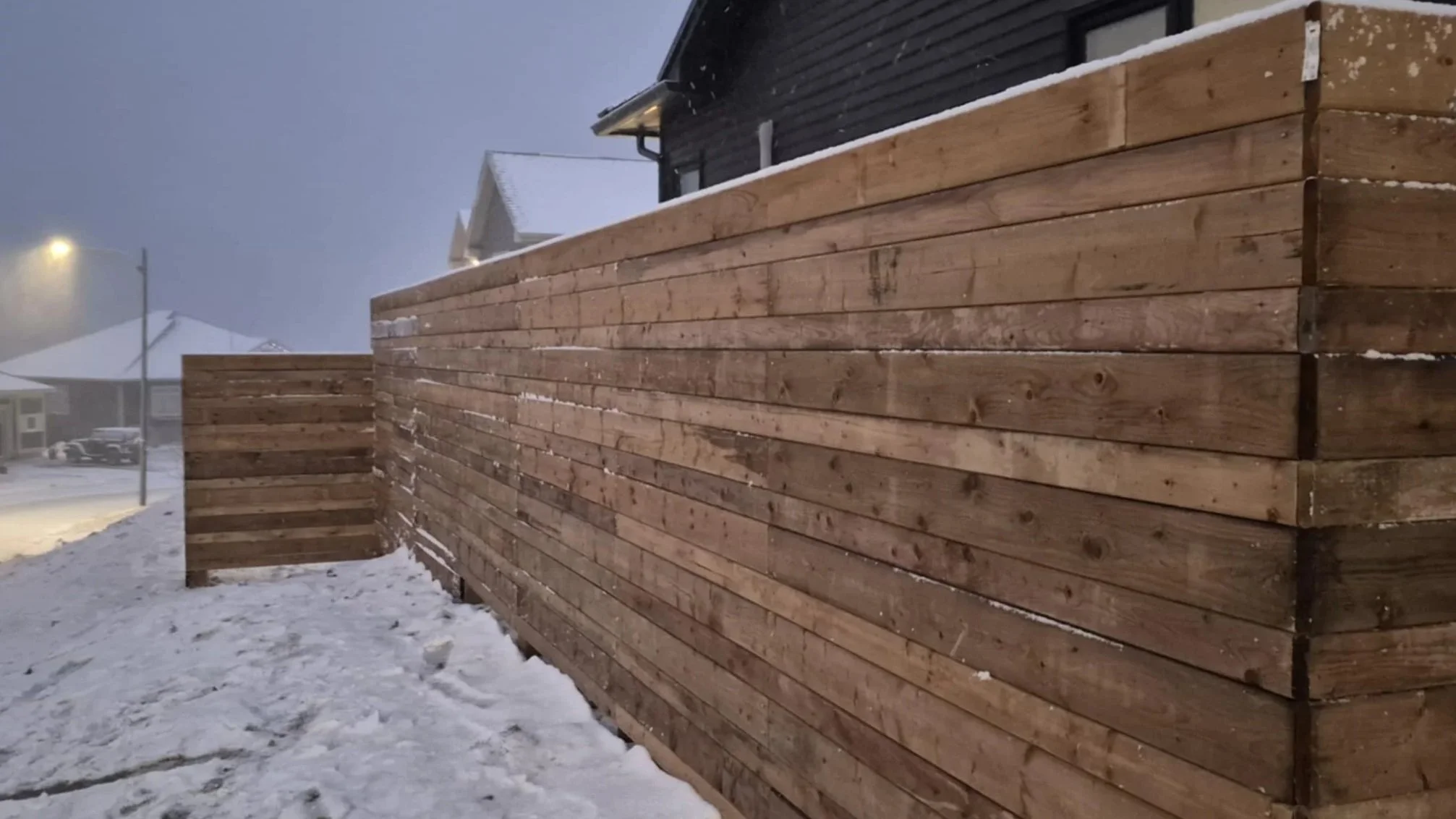 Wooden privacy fence along a snowy street with houses and streetlights in the background.