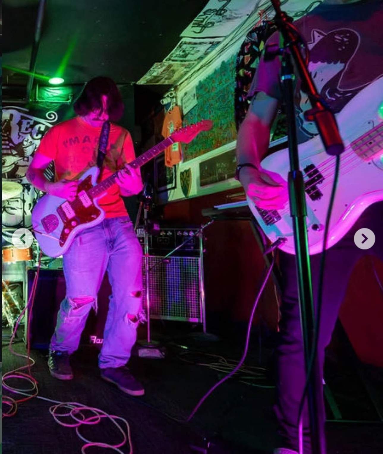 Two musicians playing guitars on stage in a dimly lit venue with colorful neon lights.