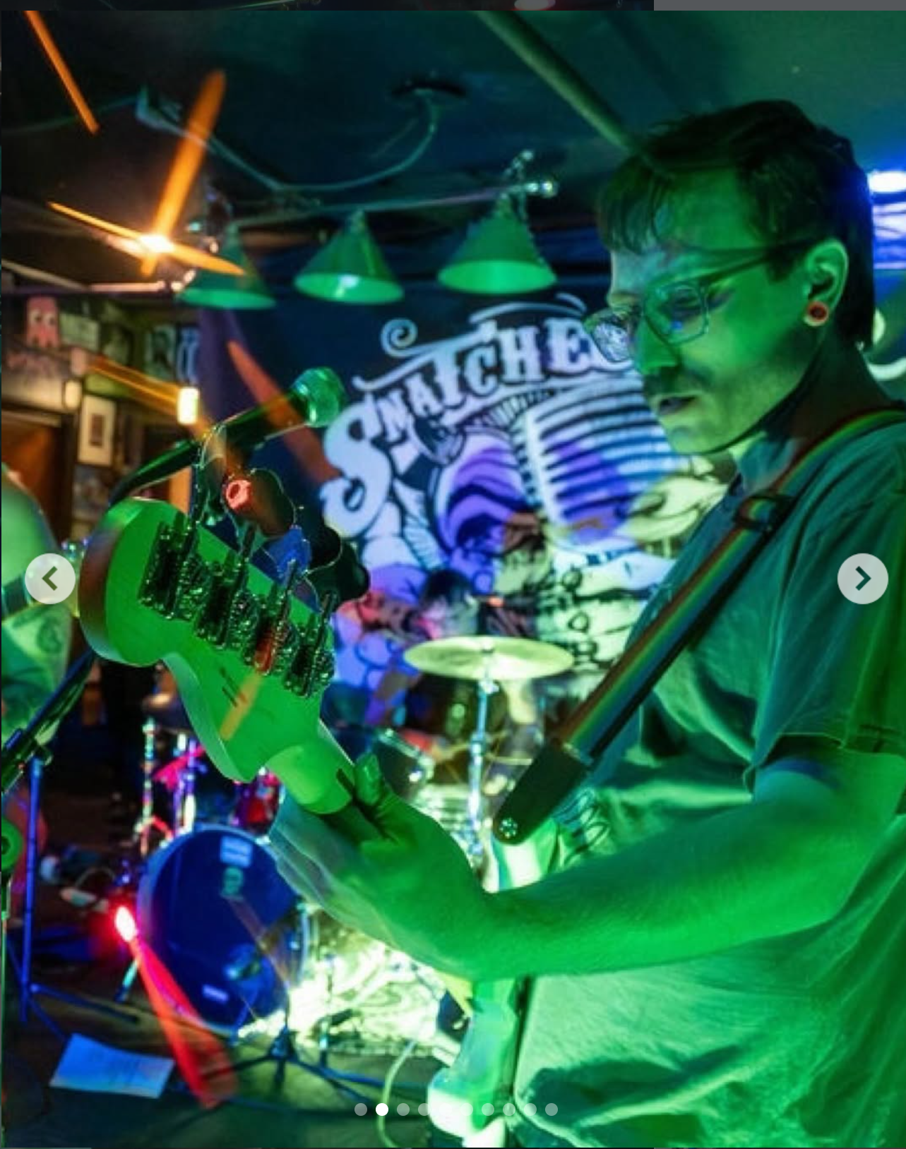 A musician playing an electric guitar on stage with a colorful backdrop, neon lighting, and the partially visible word 'SNACHE' in the background.