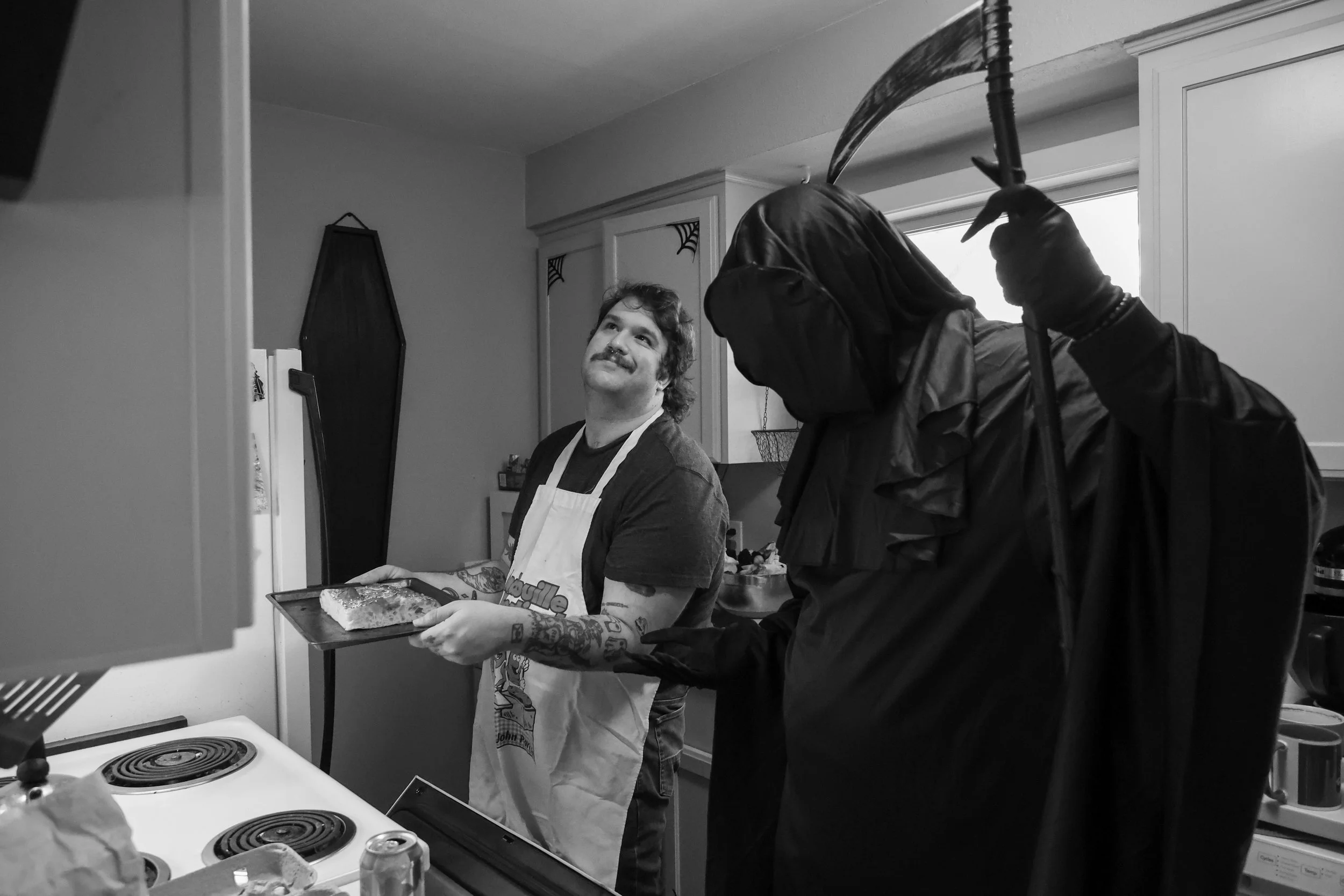 A man in an apron holding a baking tray smiling in a kitchen, with a person dressed as a grim reaper figure standing nearby.
