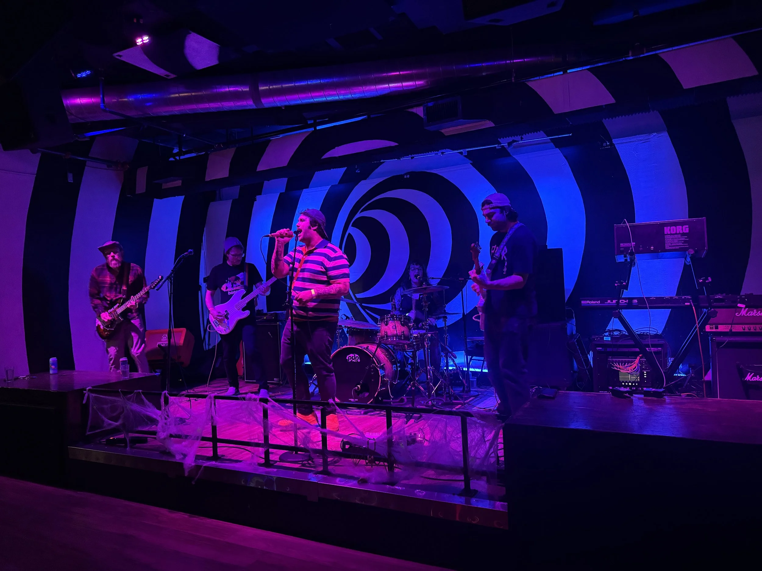 Band performing on a stage with spiral background, purple and blue lighting, with guitars, drums, keyboard, and singer in striped shirt, Halloween decorations like spider webs on the front of the stage