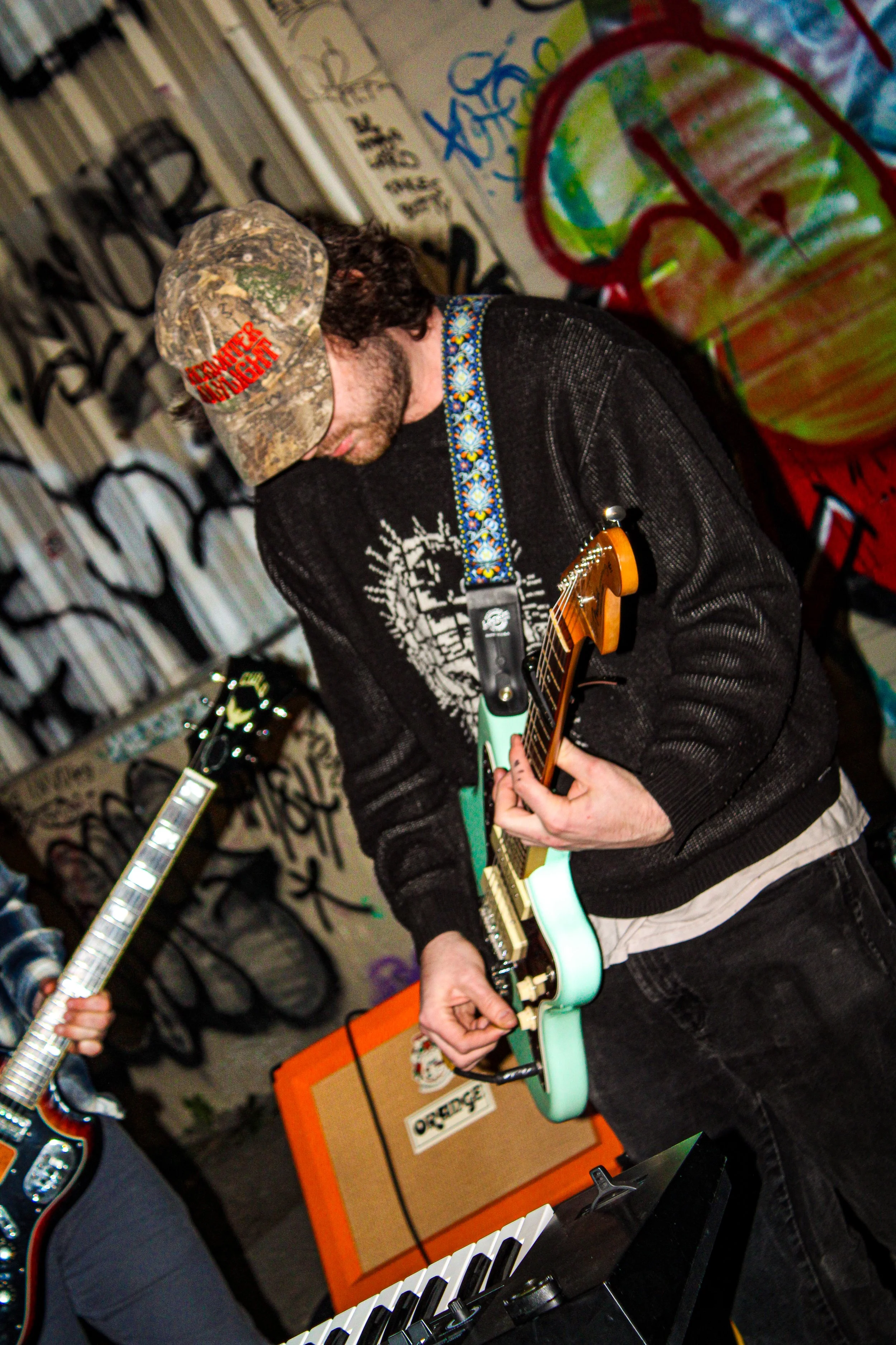 A man playing an electric guitar with a floral strap, wearing a cap and hoodie, in front of graffiti-covered walls.