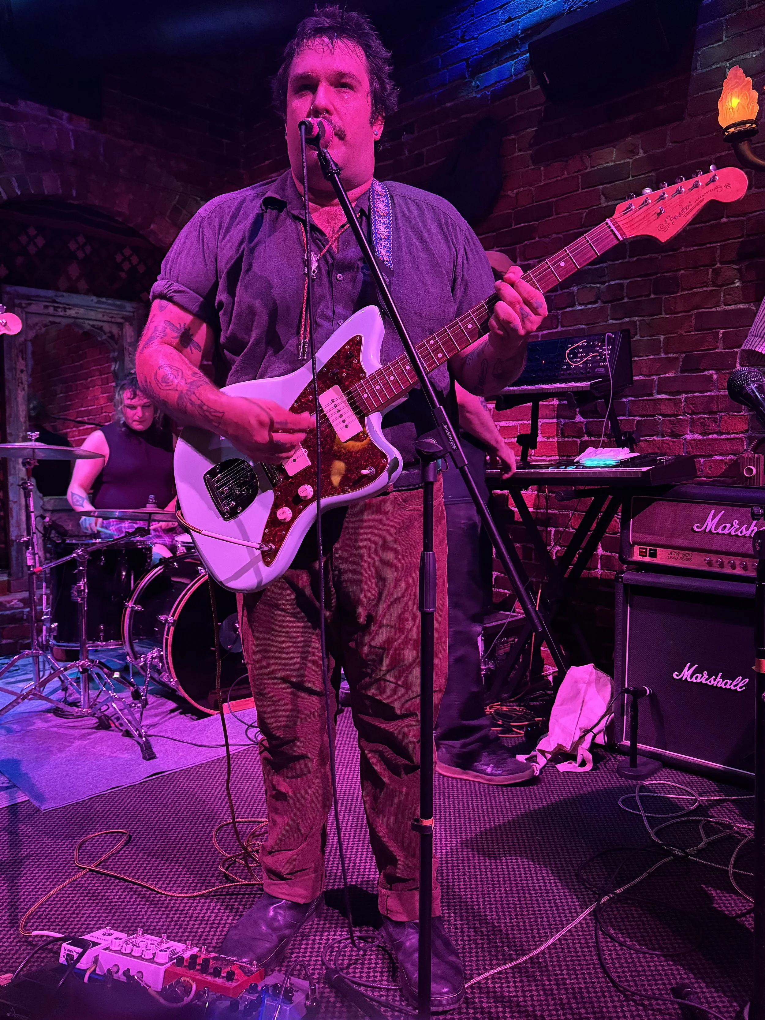 A man playing an electric guitar and singing into a microphone on stage, with a woman playing drums in the background, in a dimly lit venue with brick walls and purple and blue lighting.