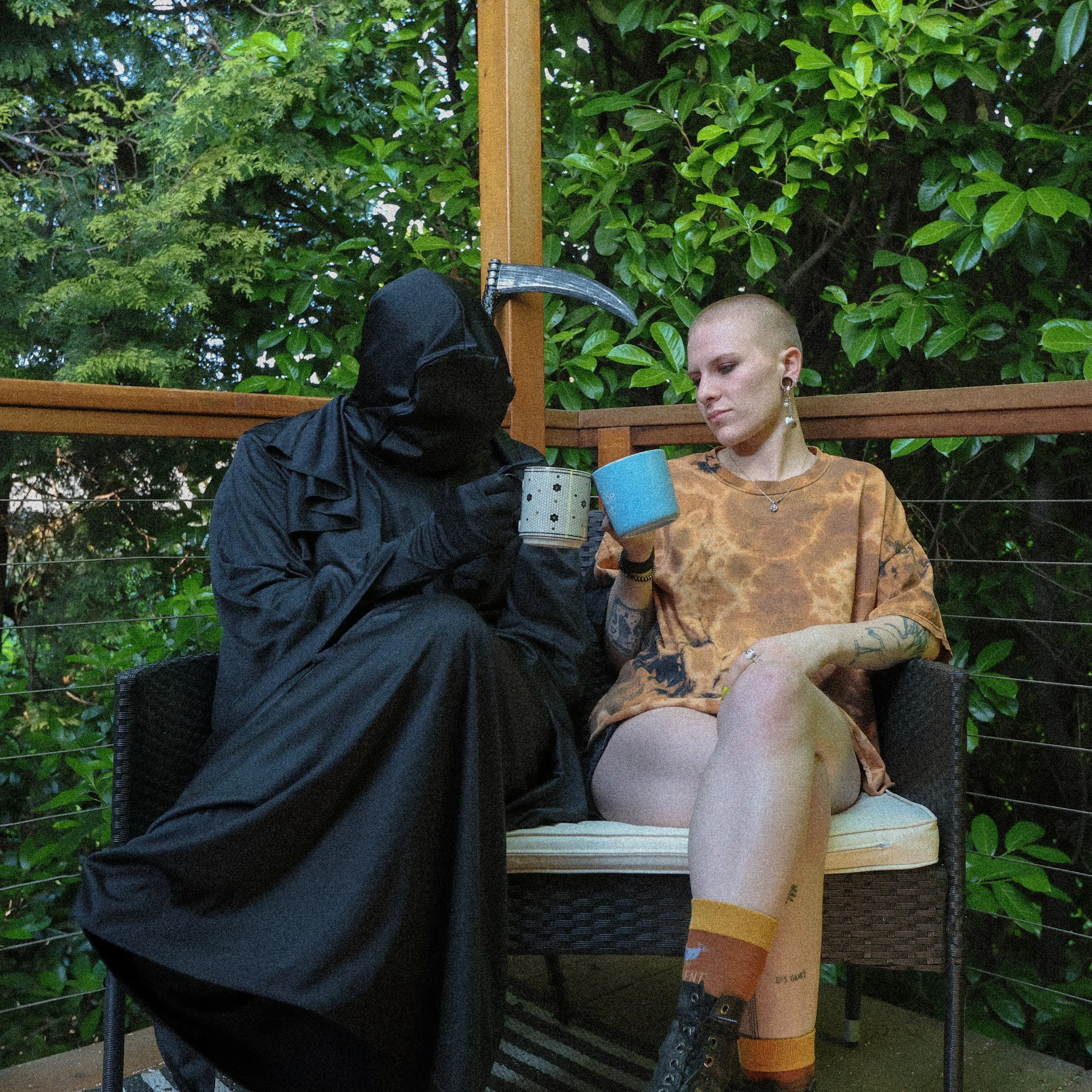 Person dressed as Grim Reaper handing a mug to a young woman with shaved head and tattoos sitting on a porch with greenery in the background.