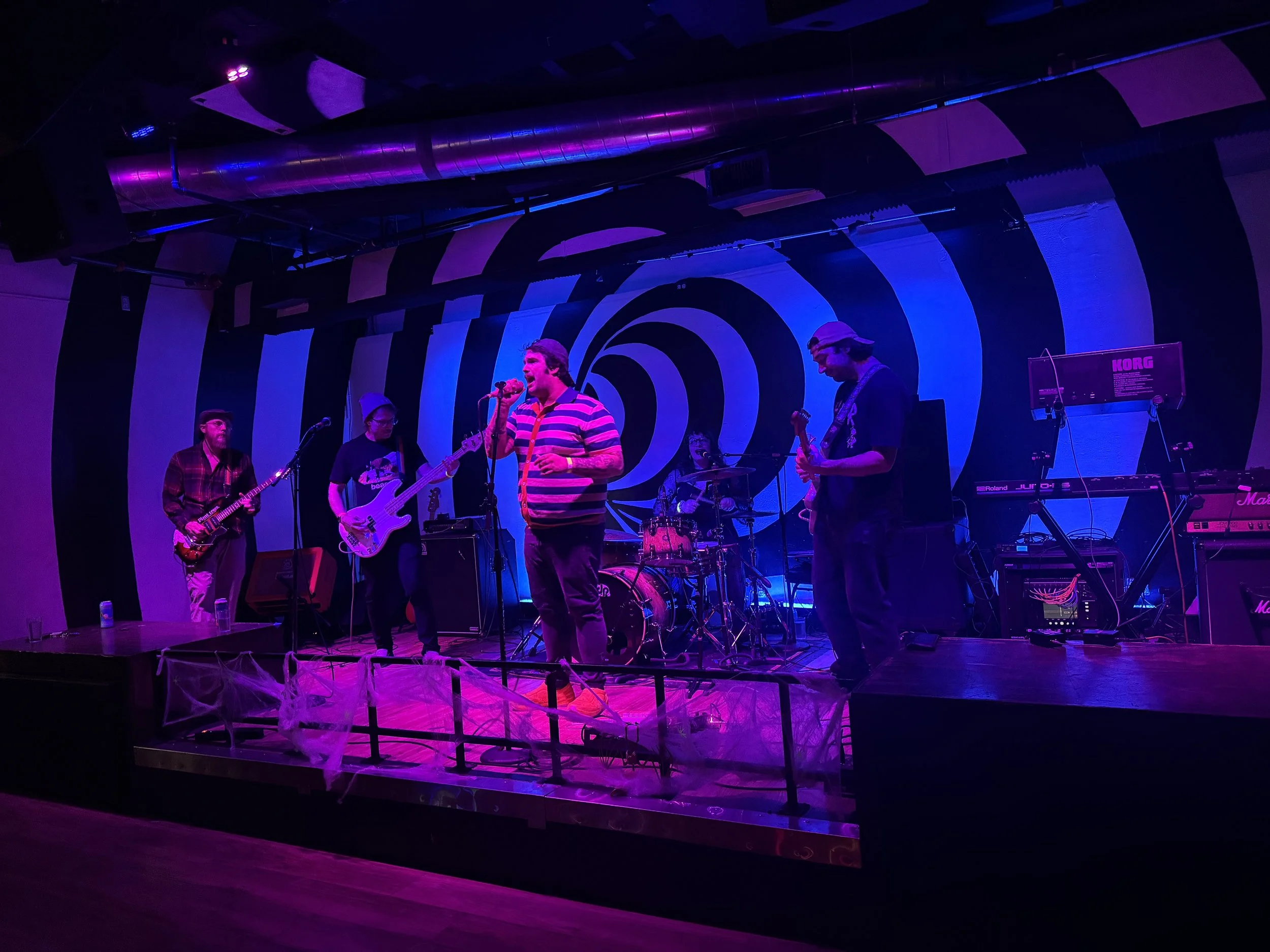 A band performs on a stage with a black and white spiral mural backdrop, illuminated by purple and blue stage lights, with members playing guitars, drums, and singing.