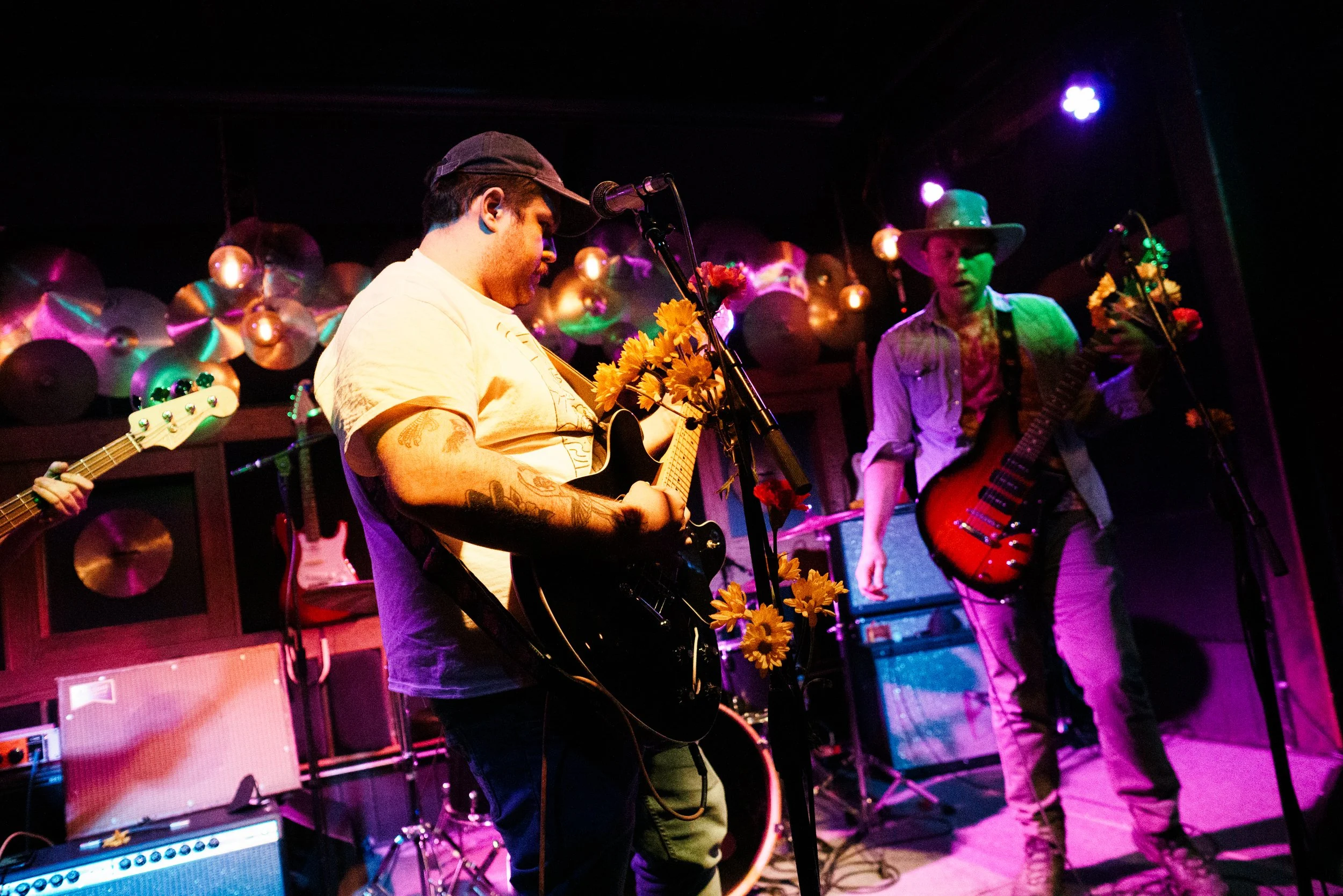 Musicians perform on stage with guitars, microphone, and floral decorations, illuminated by colorful stage lights at a live concert venue.