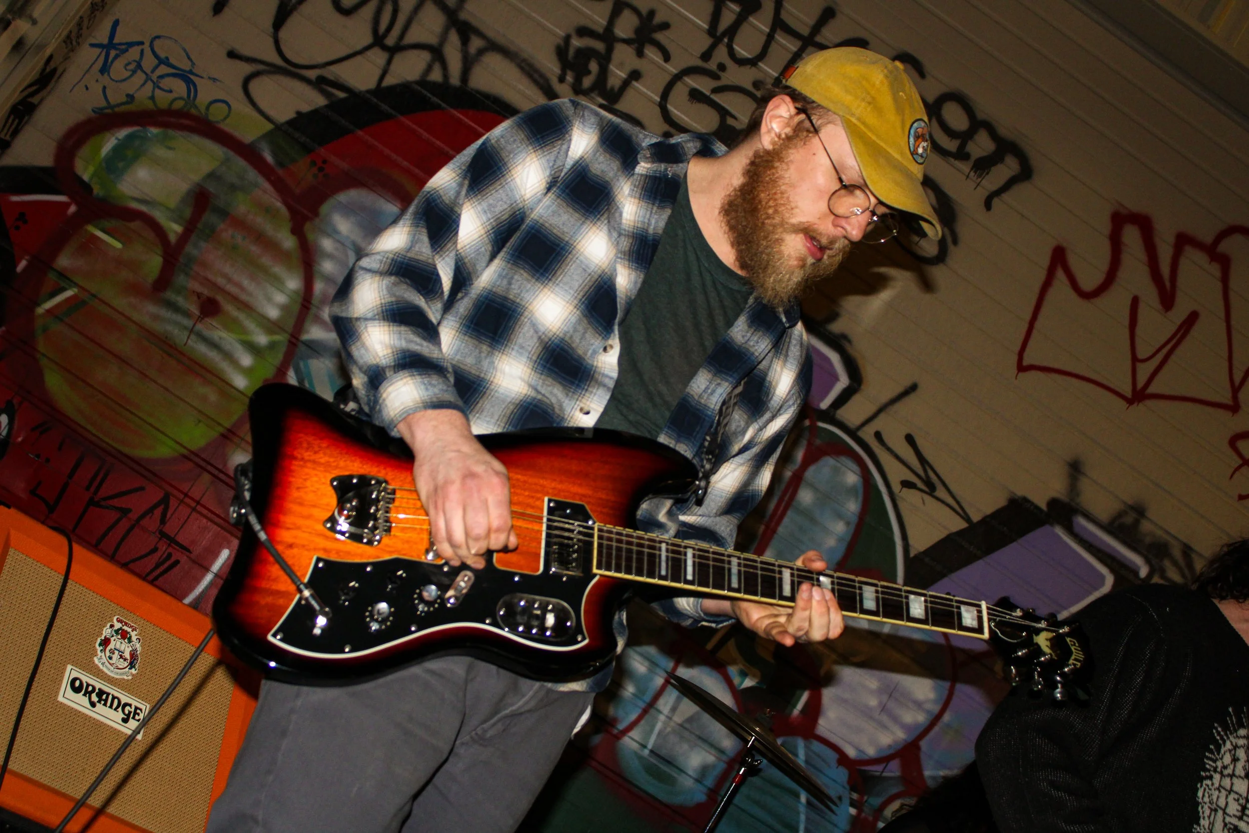 A man with glasses, a beard, and a yellow cap playing an electric guitar. He is wearing a plaid shirt and standing in front of a graffiti-covered wall.