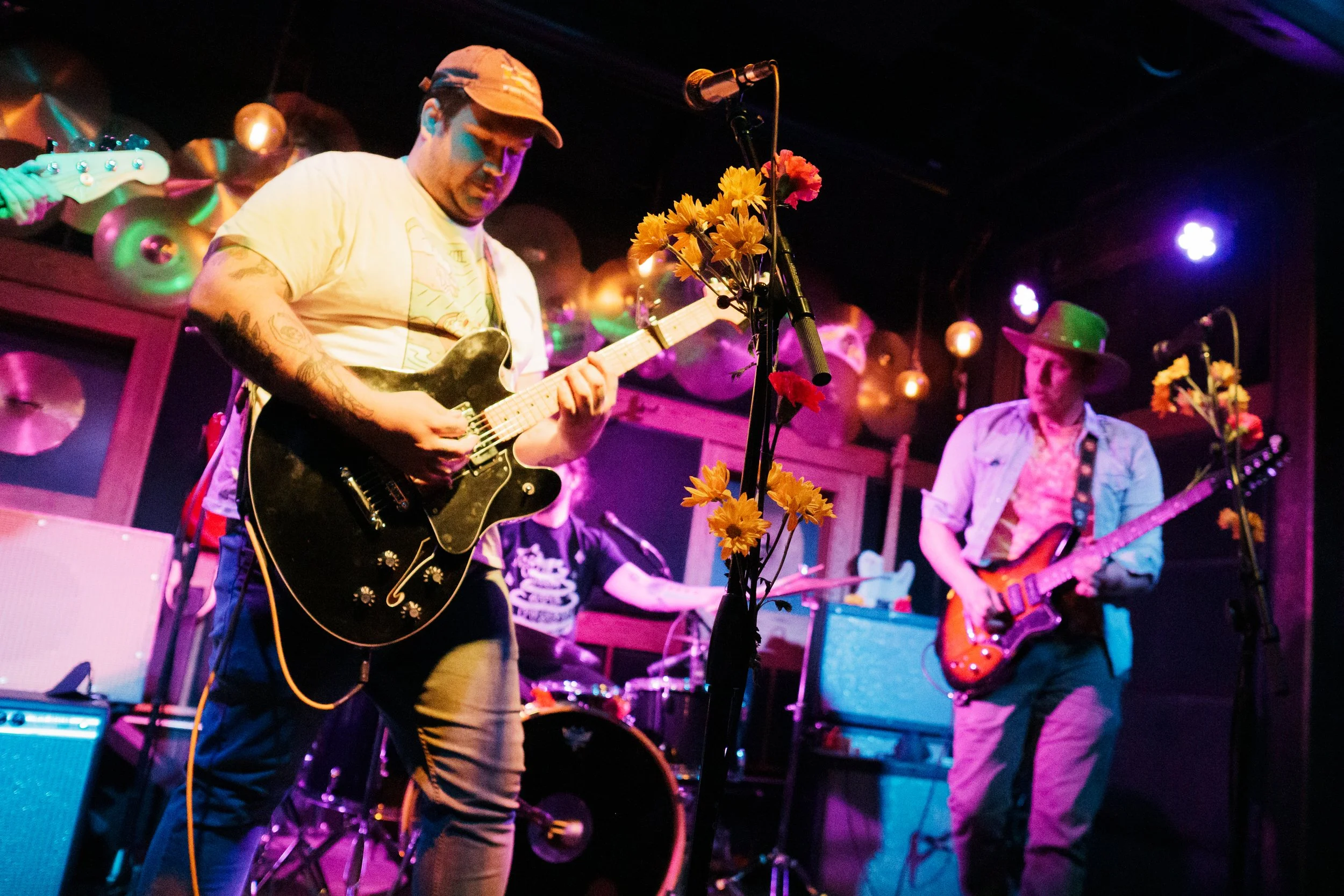 Band performing on stage with two guitarists and a drummer, decorated with yellow flowers in the foreground, colorful stage lighting in the background.