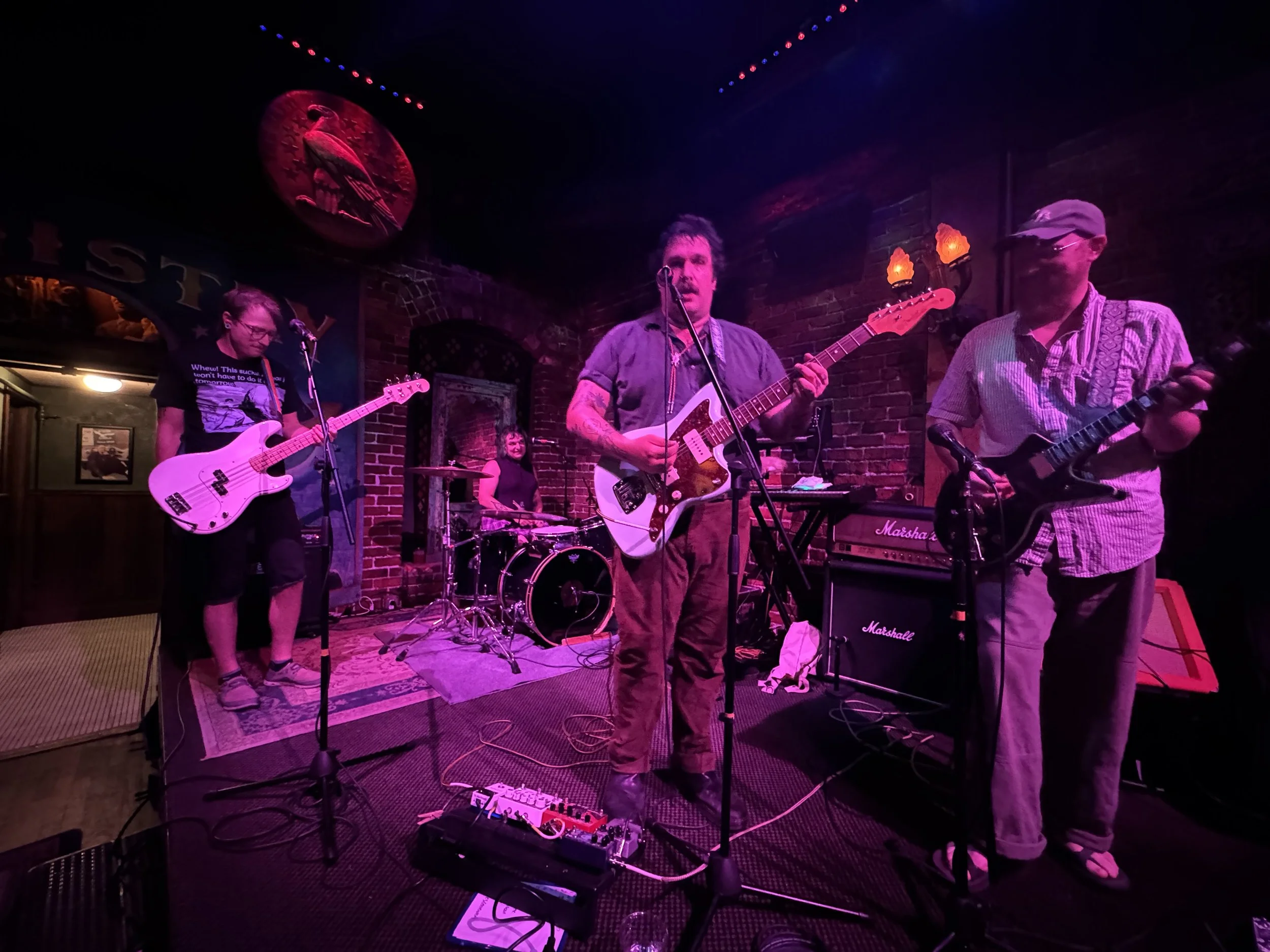 A four-piece band performing on a small stage in a dimly lit bar or club. The drummer is in the background, while three guitarists/vocalists are in the foreground, one with a bass guitar, another with an electric guitar, and the third with an electri