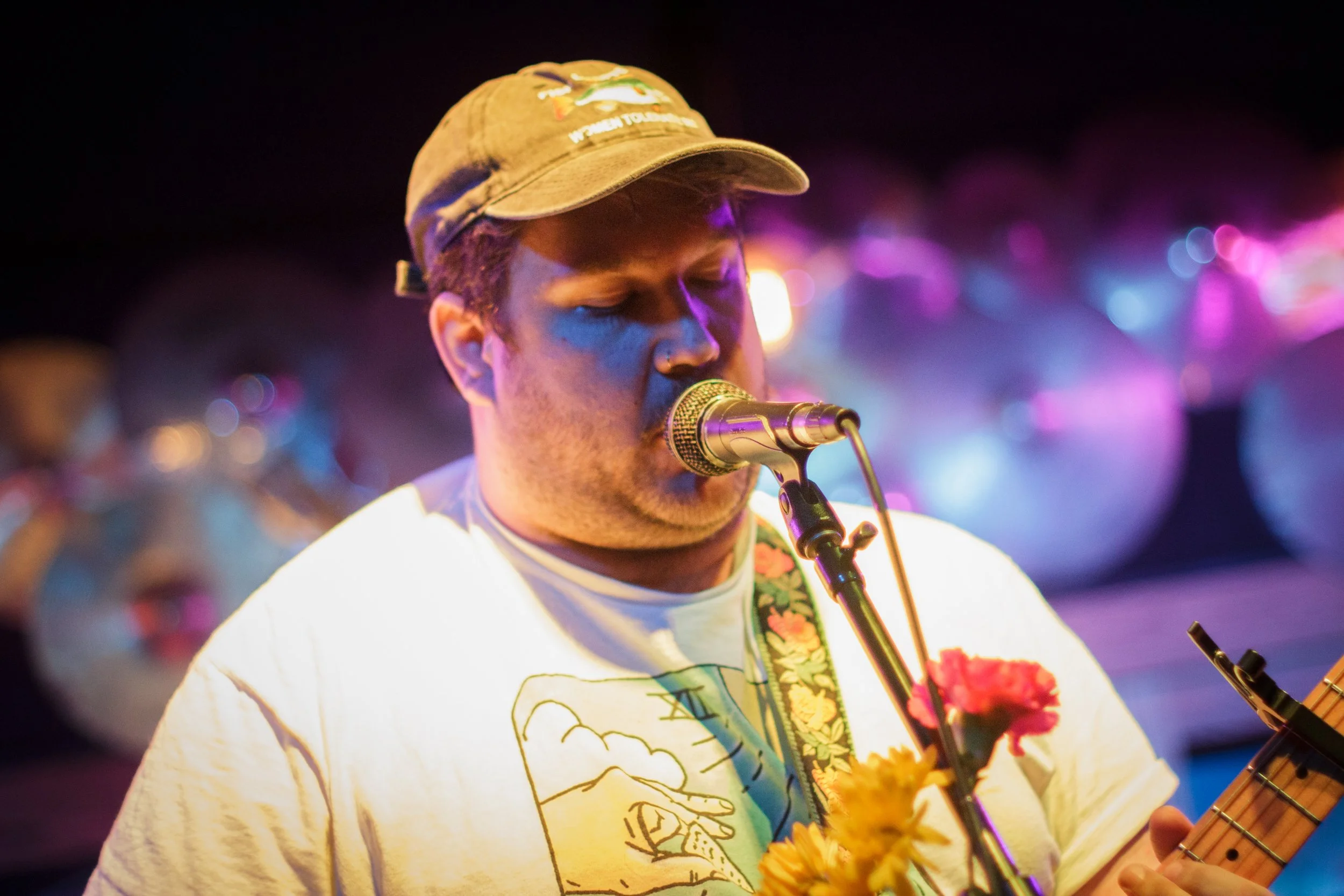 A man singing into a microphone with his eyes closed, wearing a baseball cap and a t-shirt, with colorful flowers on a guitar strap, surrounded by blurred colorful lights in the background.