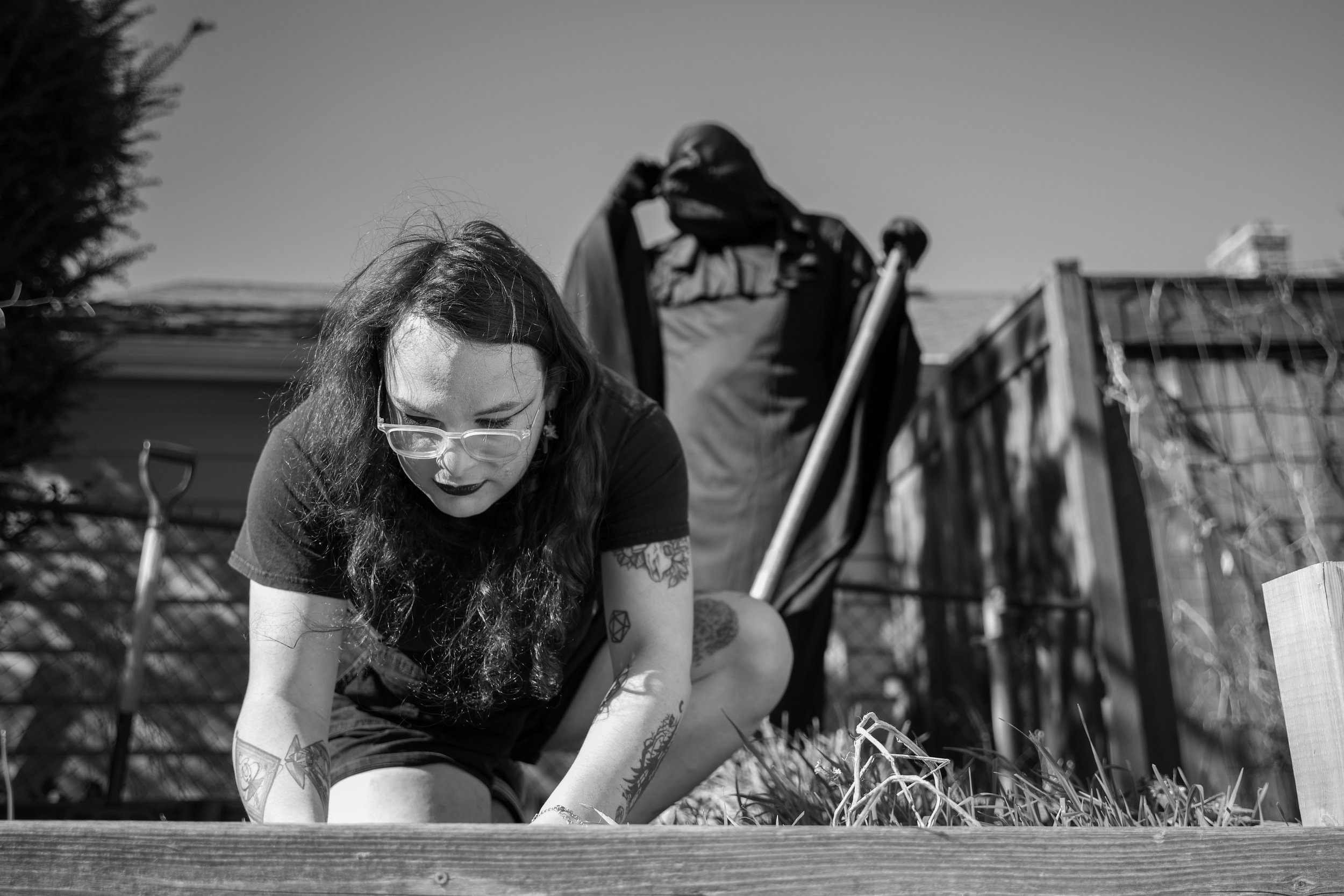 A woman with glasses and tattoos planting or gardening outside with a shovel in the background.