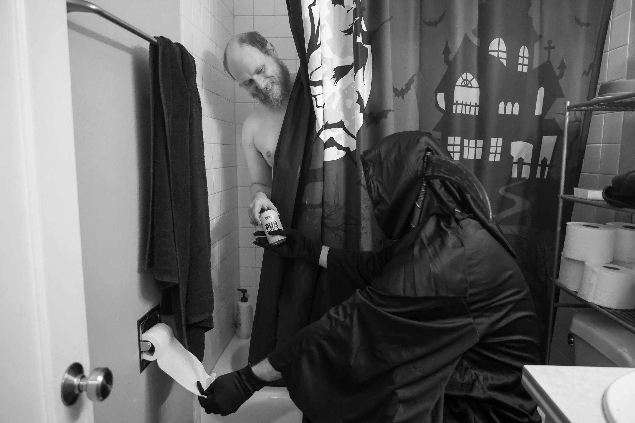 A man with a beard and no shirt smiling in a bathroom while another person in a black hooded outfit and gloves, holding cleaning supplies, cleans the toilet paper roll.