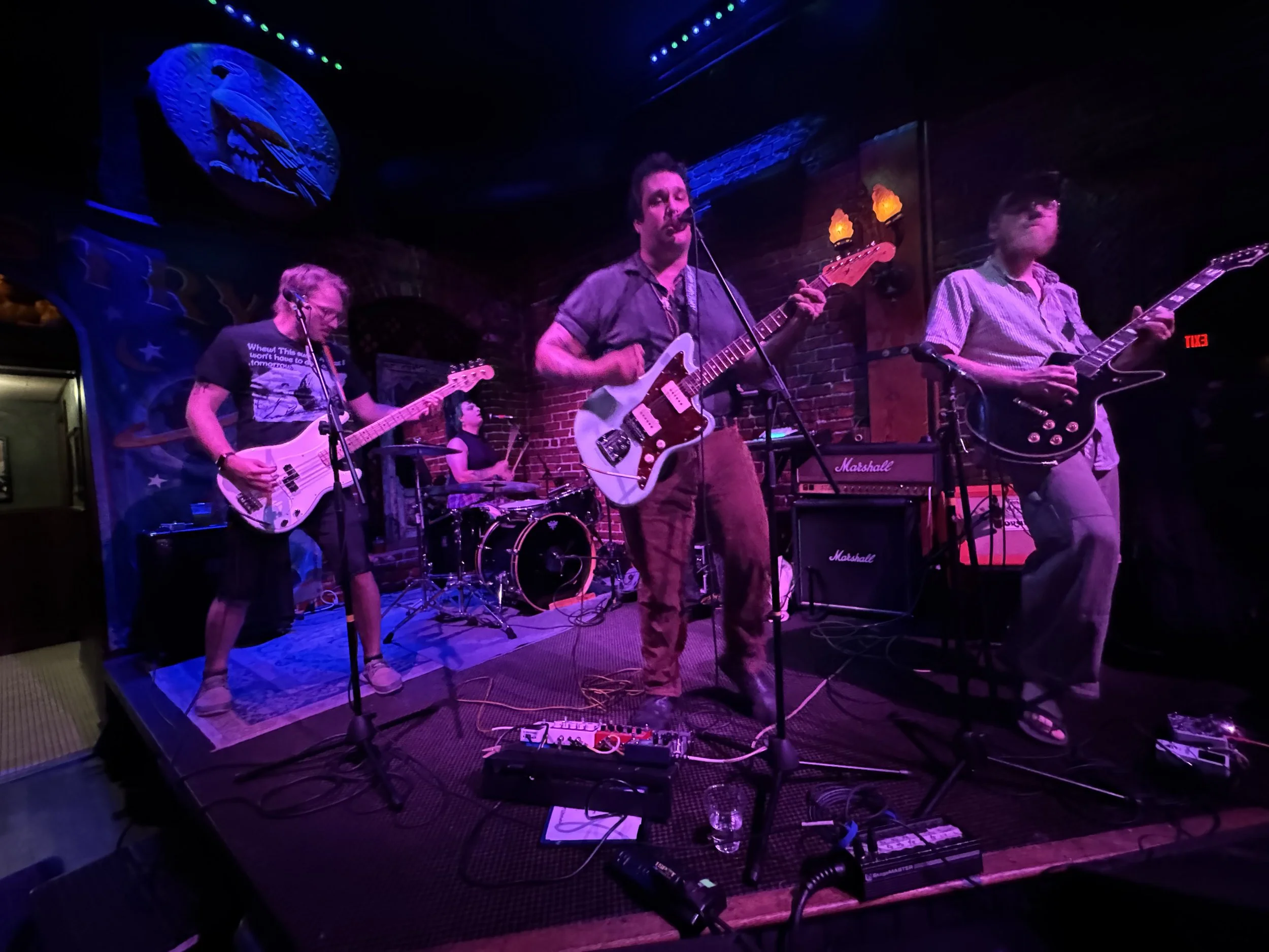 A band performing live on stage with four members playing guitars and drums at a dimly lit venue.