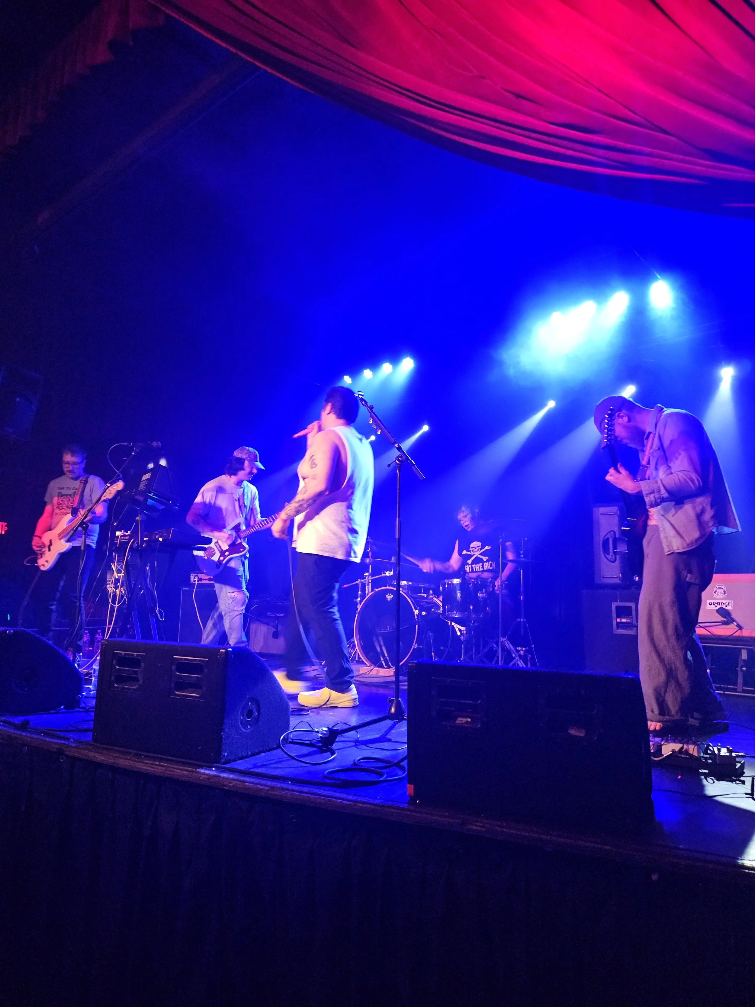 A band performing on stage with five members playing guitar, drums, and singing under colorful stage lights.