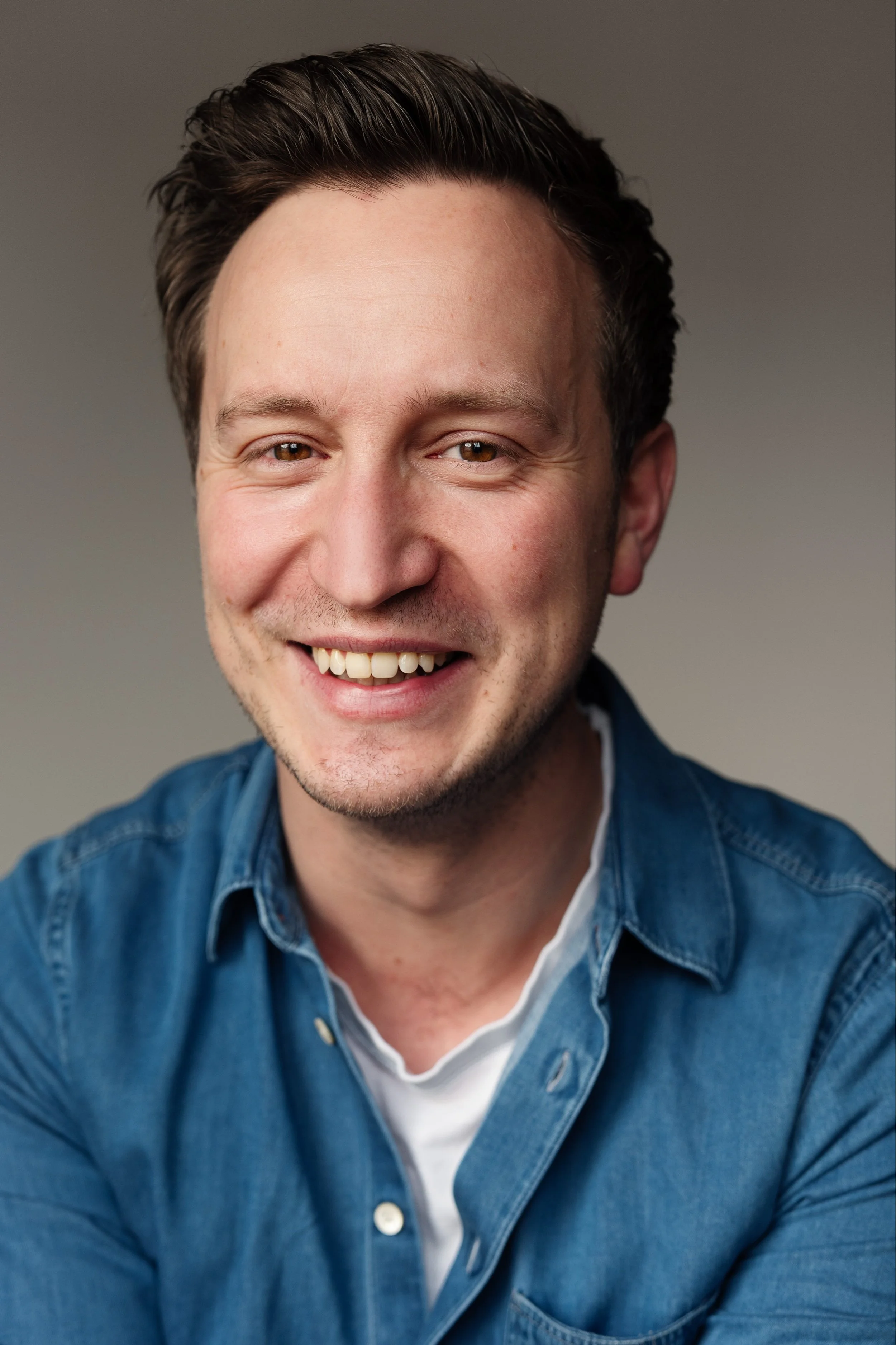 A smiling man with short dark hair, wearing a blue denim shirt and a white t-shirt, against a neutral background.