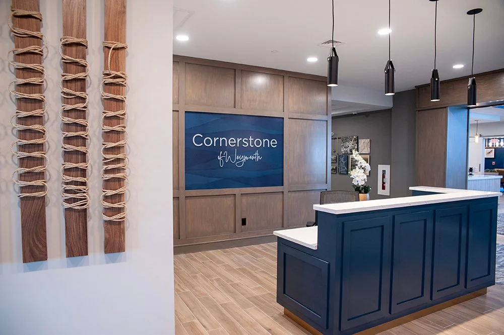 Modern office reception area with a blue front desk, wood-paneled wall, floral arrangement, and wall art, with a sign reading 'Cornerstone of Nequary'.