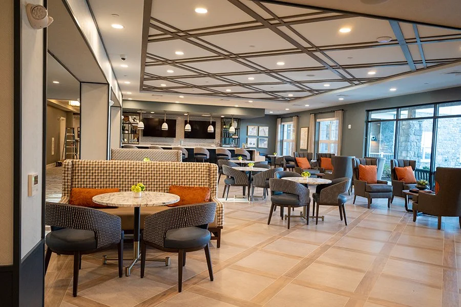 A spacious, modern lounge area with various chairs and small tables, some decorated with yellow flowers, large windows, and a bar area in the background.