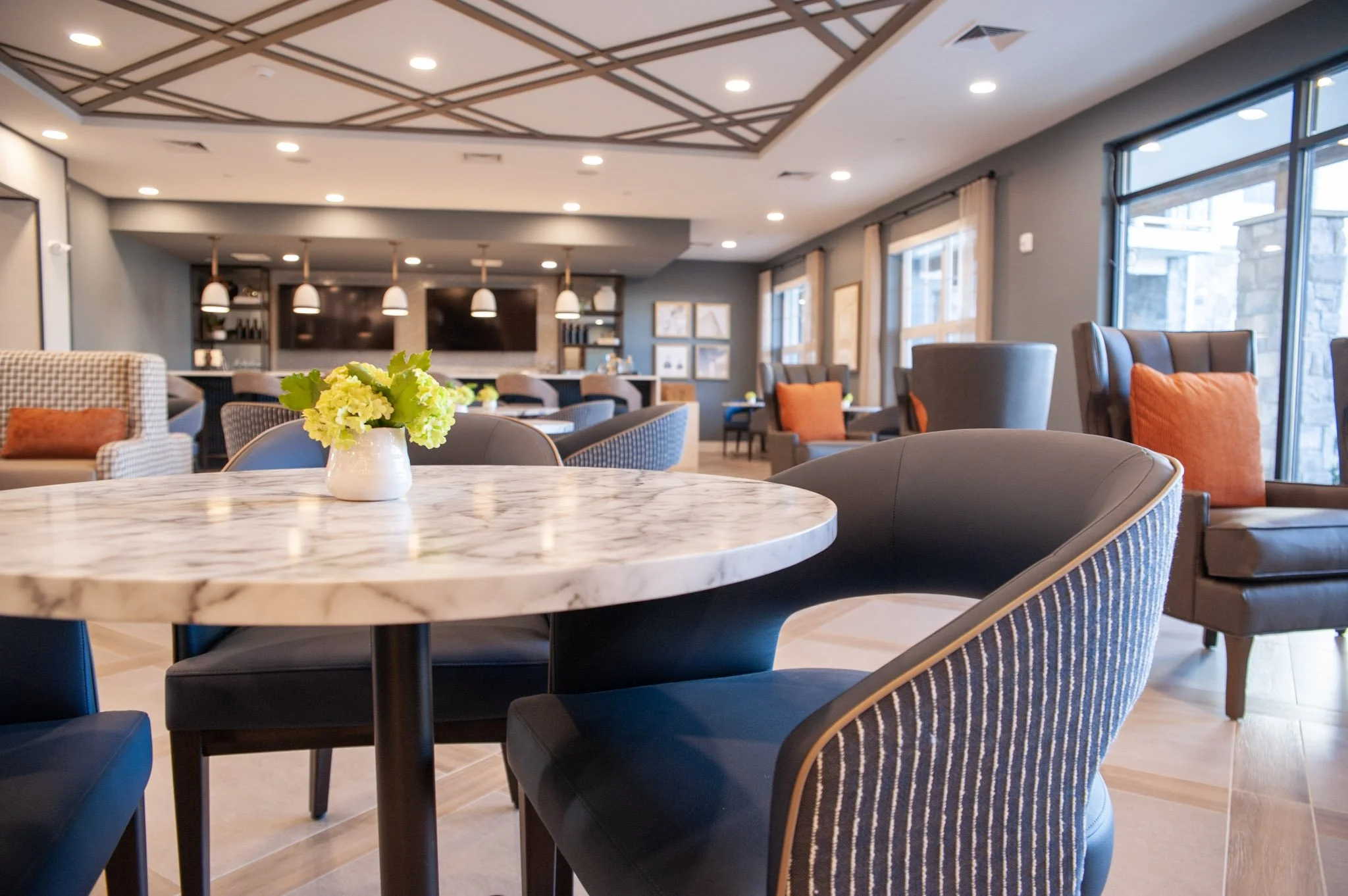 A modern, well-lit lounge with a round marble table and various armchairs, some with orange pillows. A small vase with green flowers is on the table. Large windows with curtains line one side of the room, and a flat screen TV is mounted on the wall in the background.