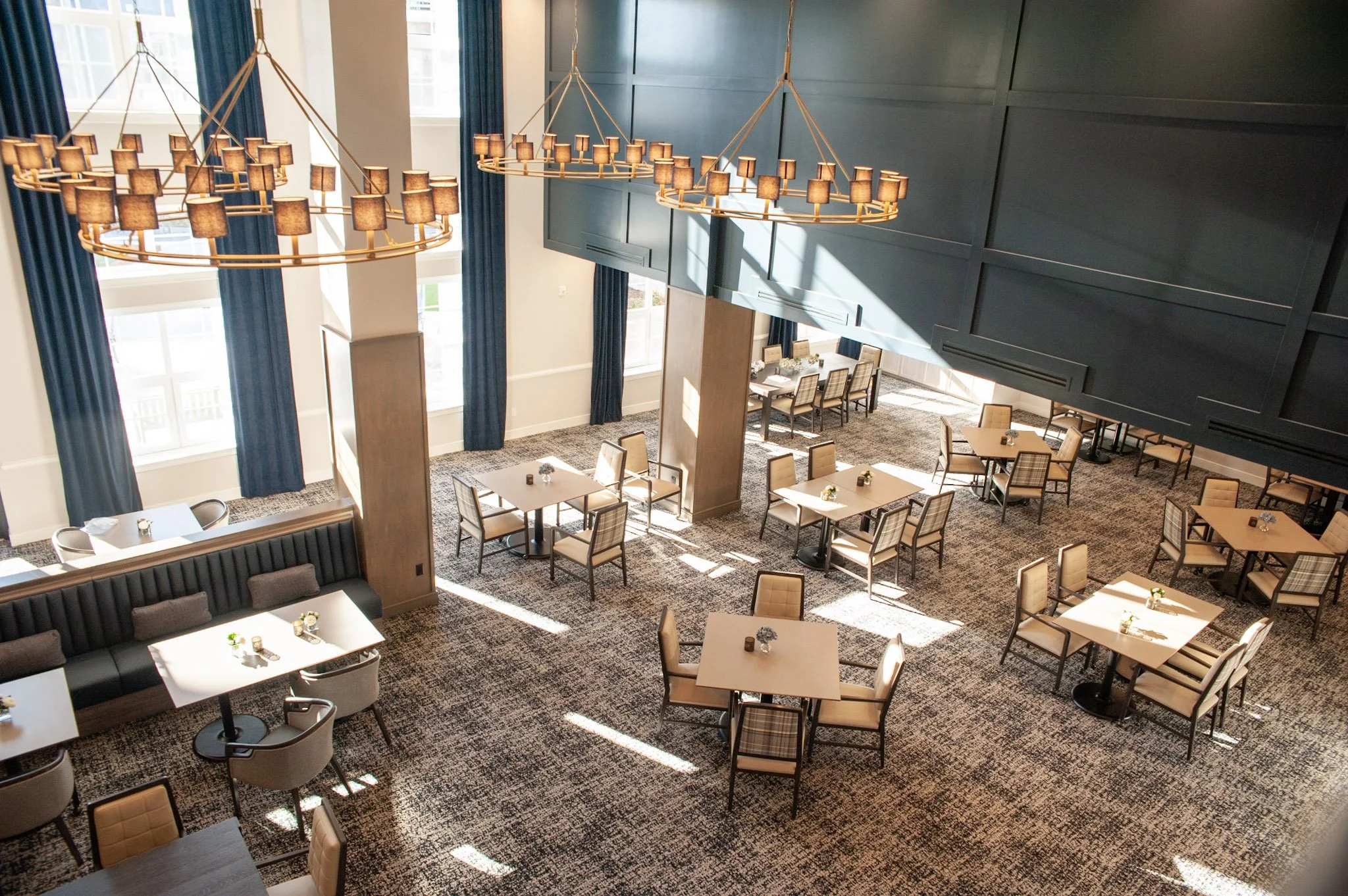 Interior of a modern restaurant or dining area with multiple square tables, beige cushioned chairs, some with plaid upholstery. Large windows with blue curtains, dark blue wall panels, and two large circular chandeliers with small shades hanging from the ceiling. Sunlight streams in, creating patches of light on the carpeted floor.