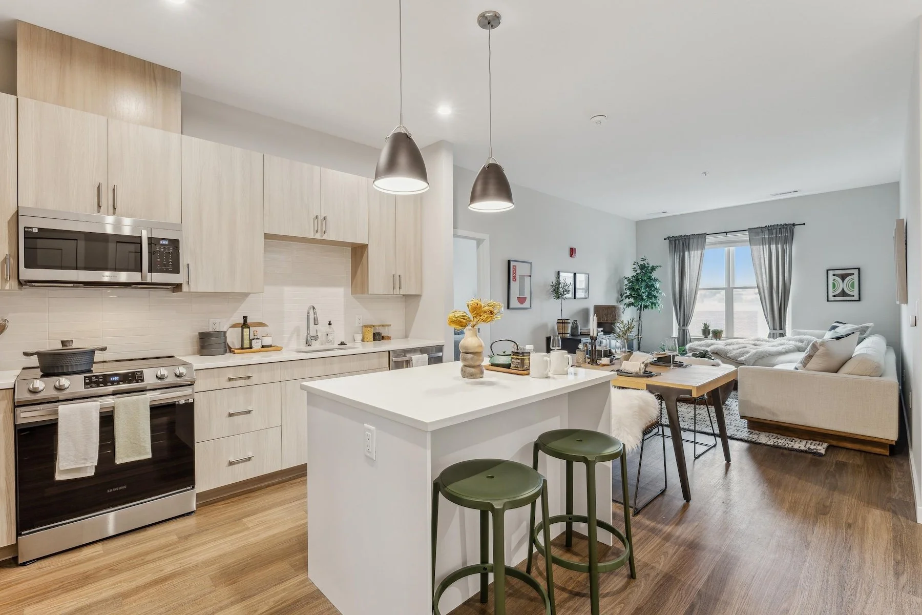 Open-concept kitchen and living room with natural light, white cabinets, a kitchen island with green stools, and a beige sofa with decorative pillows.