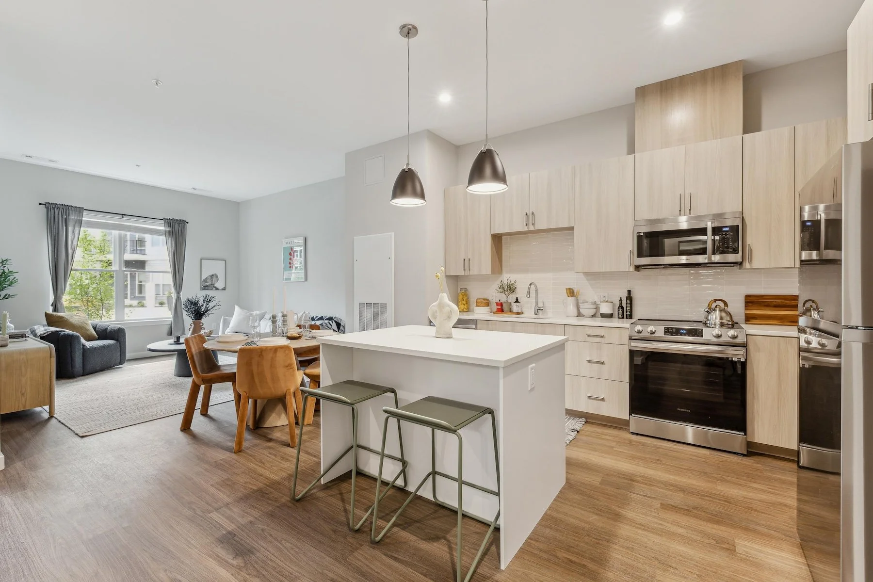 Open-concept living room and kitchen with wood flooring, light-colored cabinets, and stainless steel appliances, including a microwave, oven, and refrigerator. The living area has a window with gray curtains, a dark sofa, and a small wooden table.