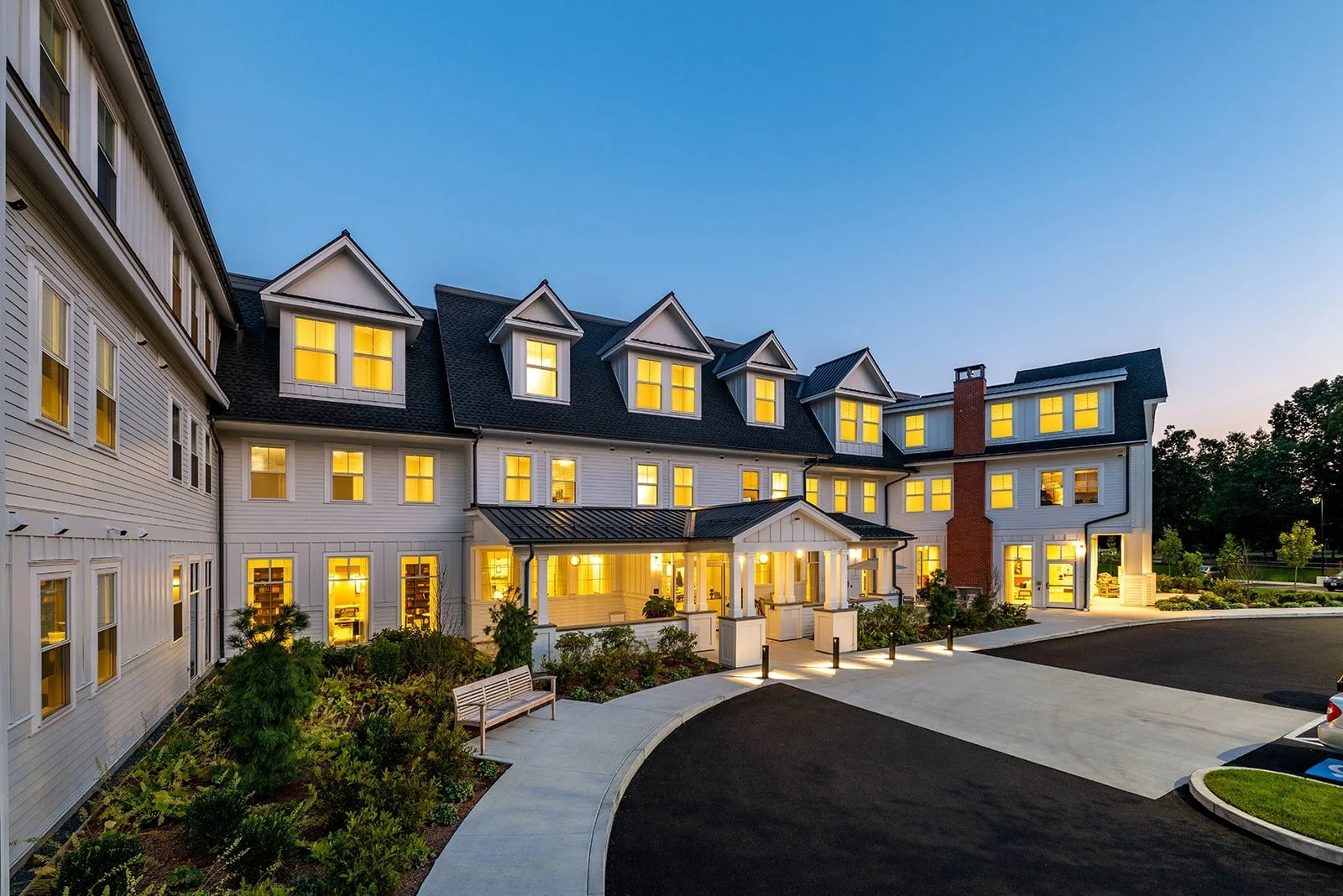 A multi-story residential building with white siding, illuminated windows, and a black roof, surrounded by a landscaped area and a parking lot at dusk.