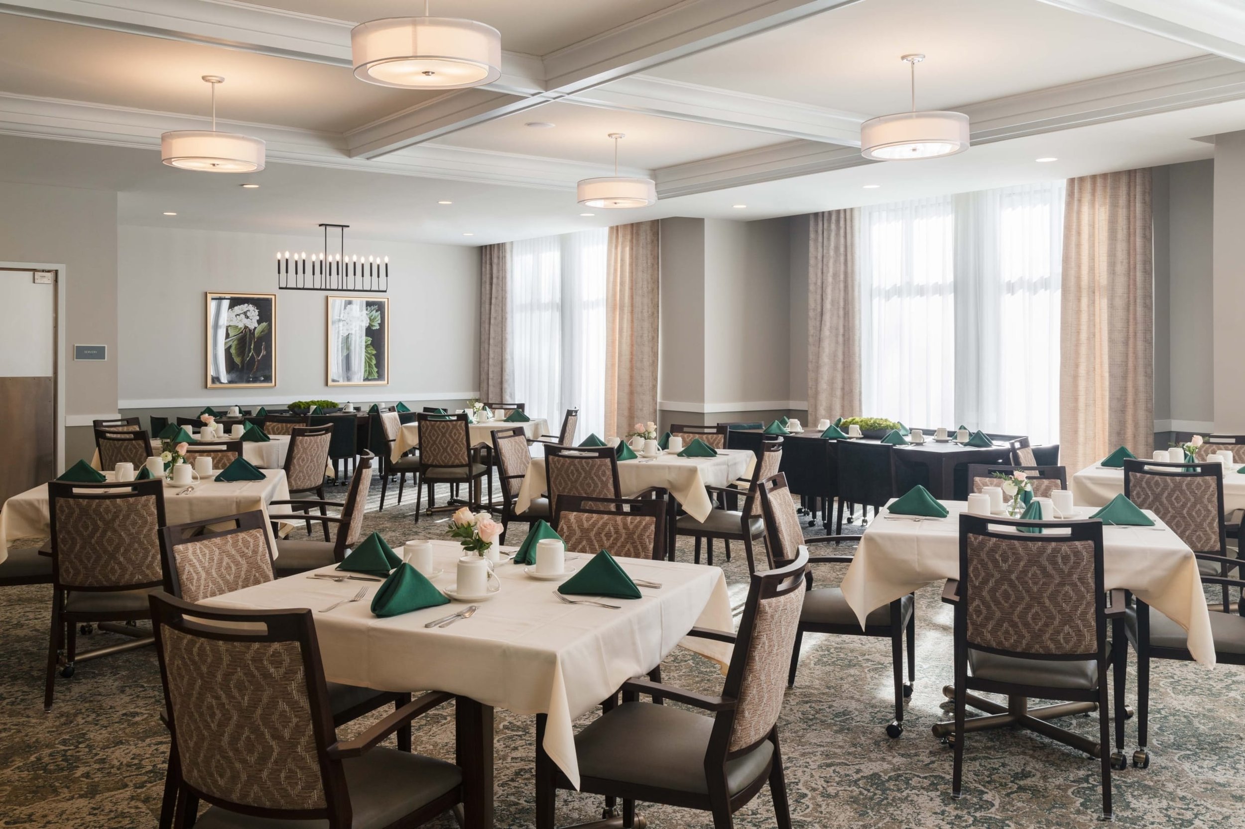 Dining room with several round tables covered with white tablecloths, set with silverware, green napkins, white coffee mugs, and small flower centerpieces. The room has large windows with sheer curtains allowing natural light, beige carpeting, and modern light fixtures on the ceiling.