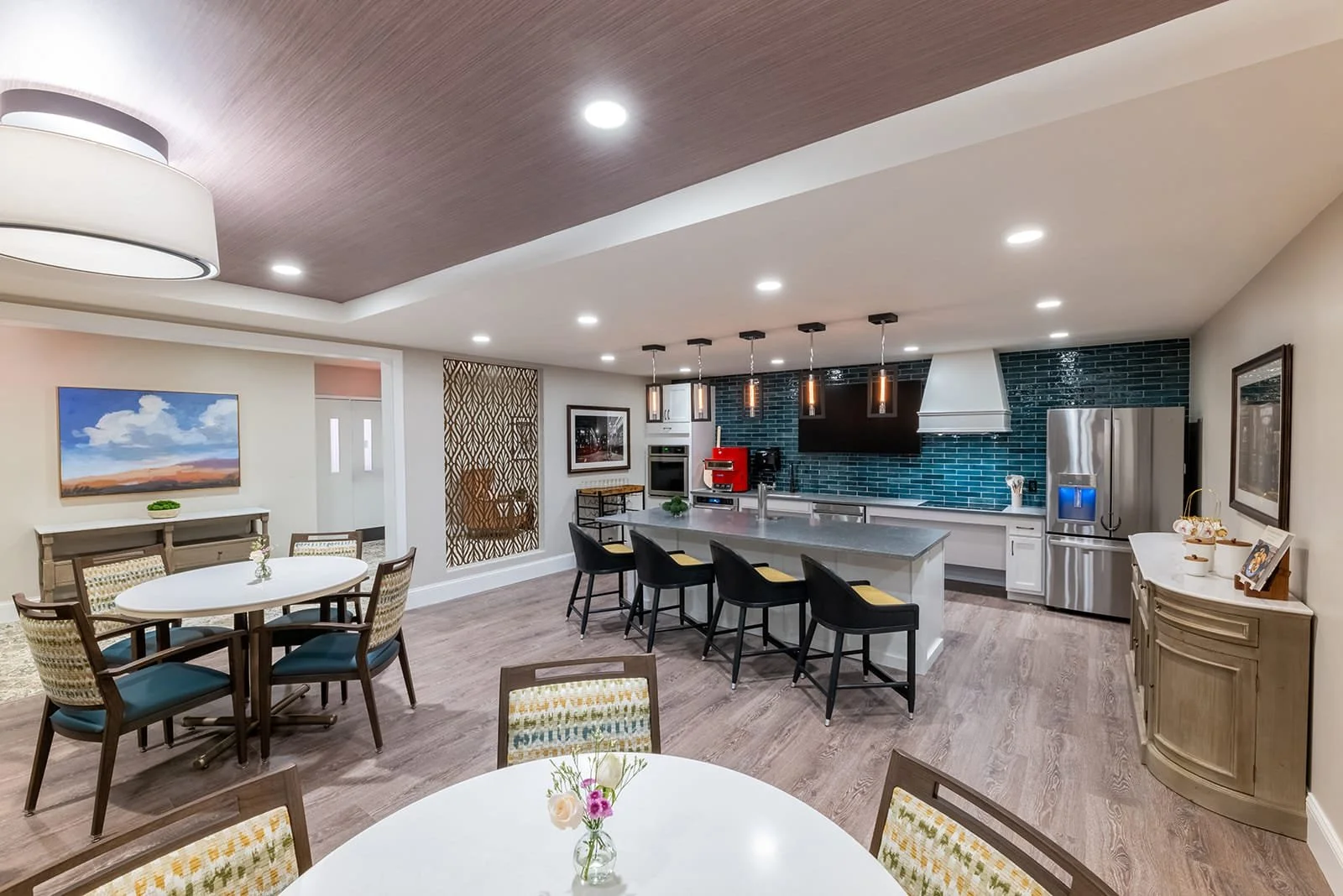 A modern, open-concept kitchen and dining area with round tables and chairs, a large kitchen island with five black chairs, blue subway tile backsplash, stainless steel refrigerator, white cabinetry, and pendant lighting