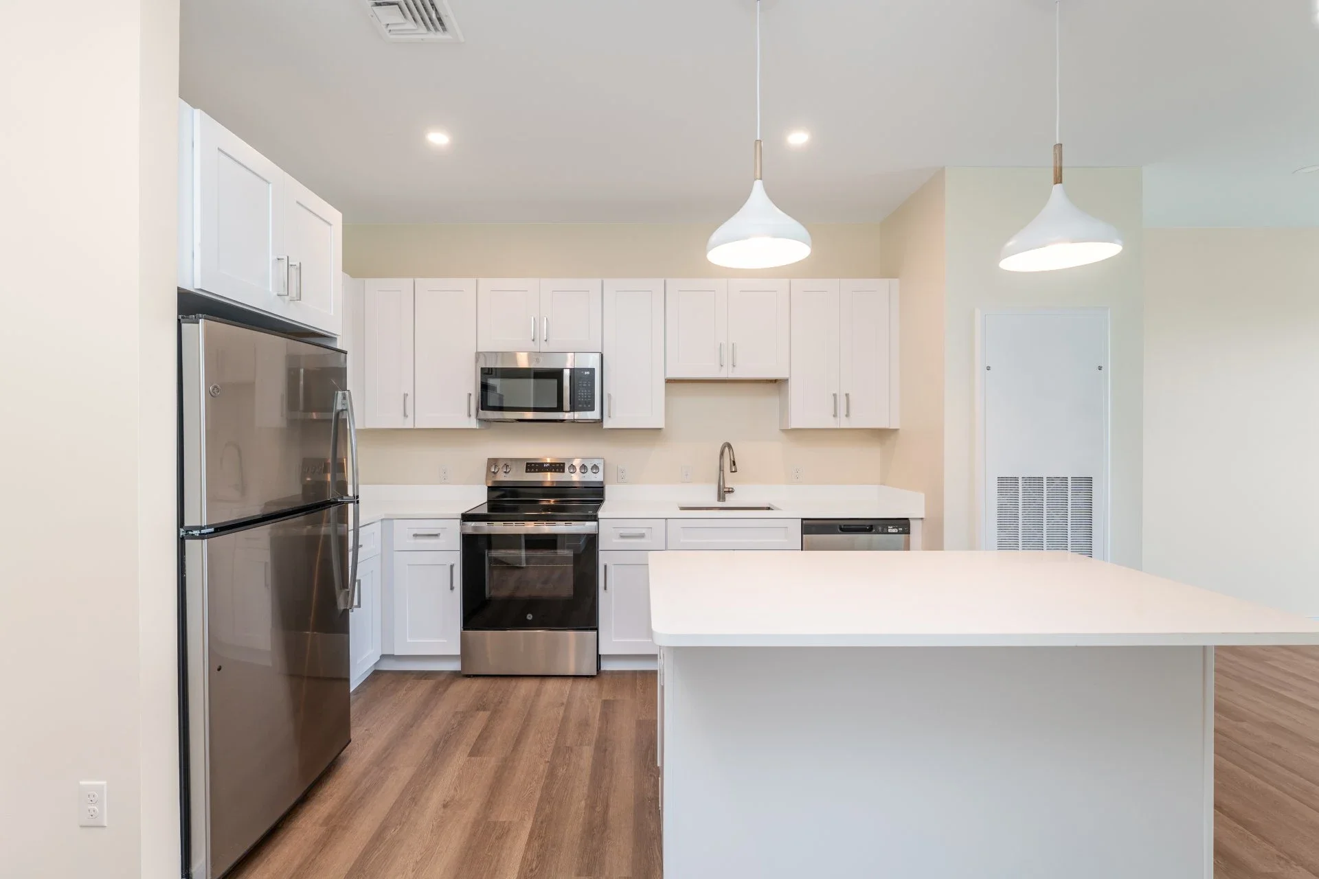 Modern kitchen with white cabinets, stainless steel refrigerator, stove, microwave, dishwasher, and a white countertop island. Two pendant lights hang from the ceiling, and there are recessed ceiling lights.