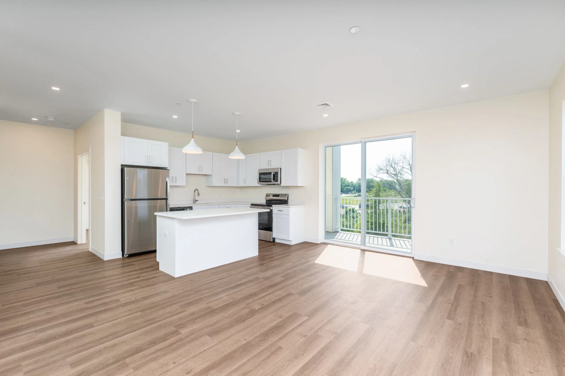 Empty open-concept kitchen with white cabinets, stainless steel refrigerator, microwave, and oven, wooden flooring, two pendant lights, and a sliding glass door leading to a balcony with views of trees and greenery.
