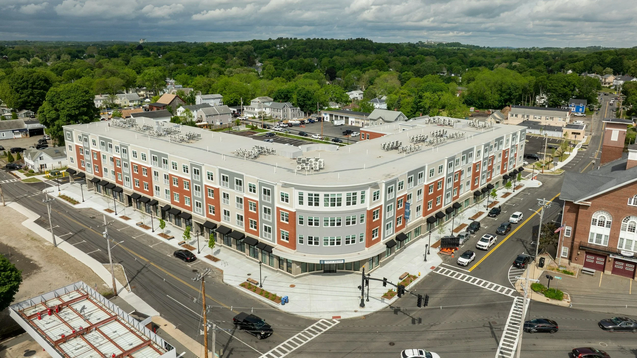 Aerial view of a modern mixed-use building at the corner of an urban intersection surrounded by residential homes and green trees.