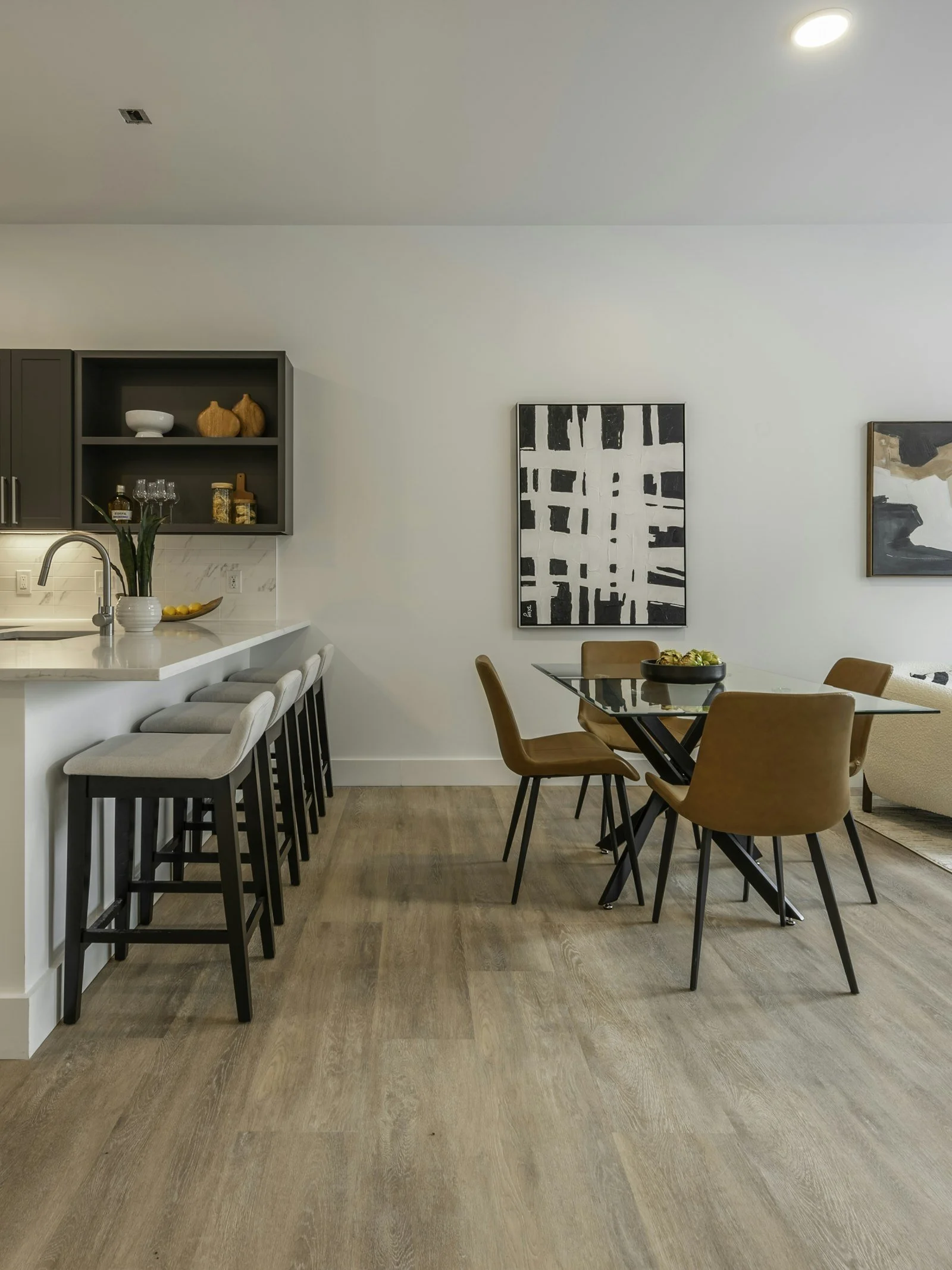 Modern kitchen and dining area featuring a white island with beige bar stools, a glass dining table with brown chairs, and abstract black and white artwork on the wall.