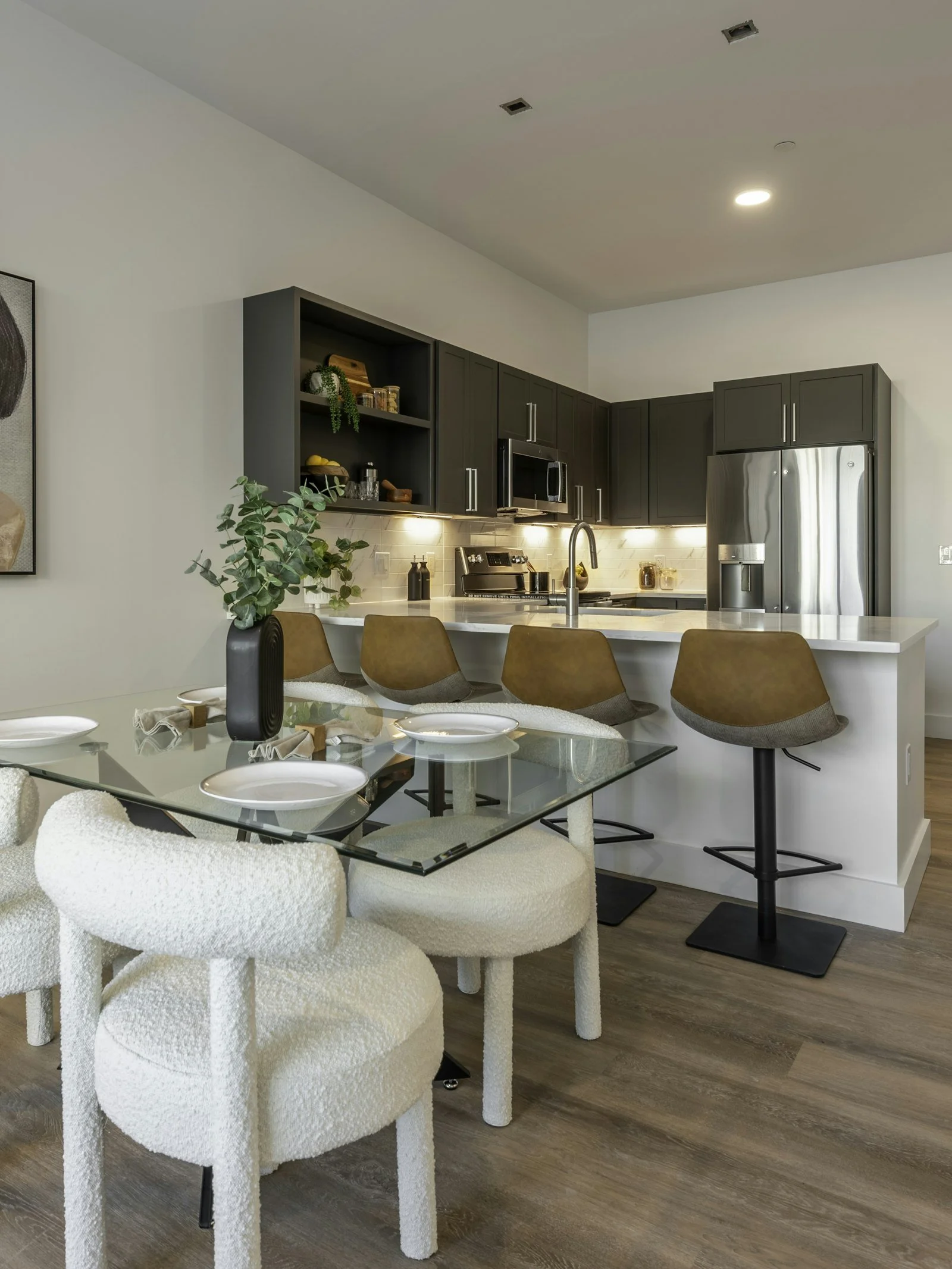 Modern kitchen with gray cabinetry, stainless steel appliances, a white countertop, and a dining table with plush white chairs. There are brown barstools at the counter and a potted plant on the table.