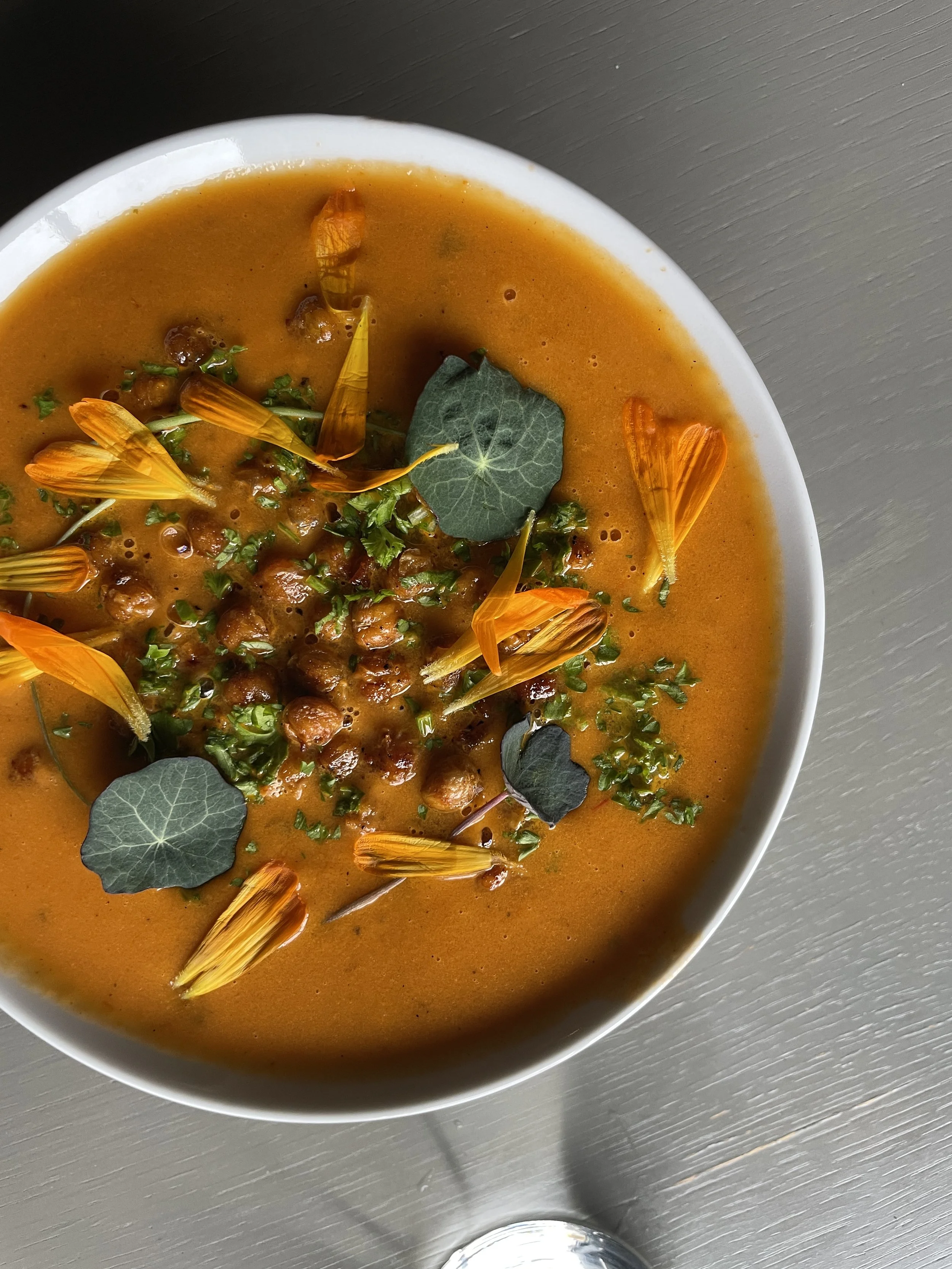 A bowl of orange-colored soup garnished with green herbs, edible flowers, and lemon balm leaves.