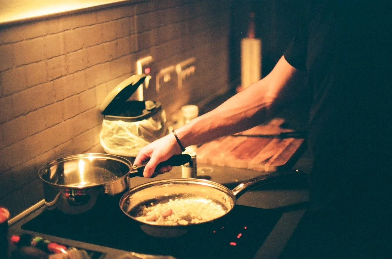 Person cooking on a stovetop with a frying pan and a pot in a kitchen with brick walls.