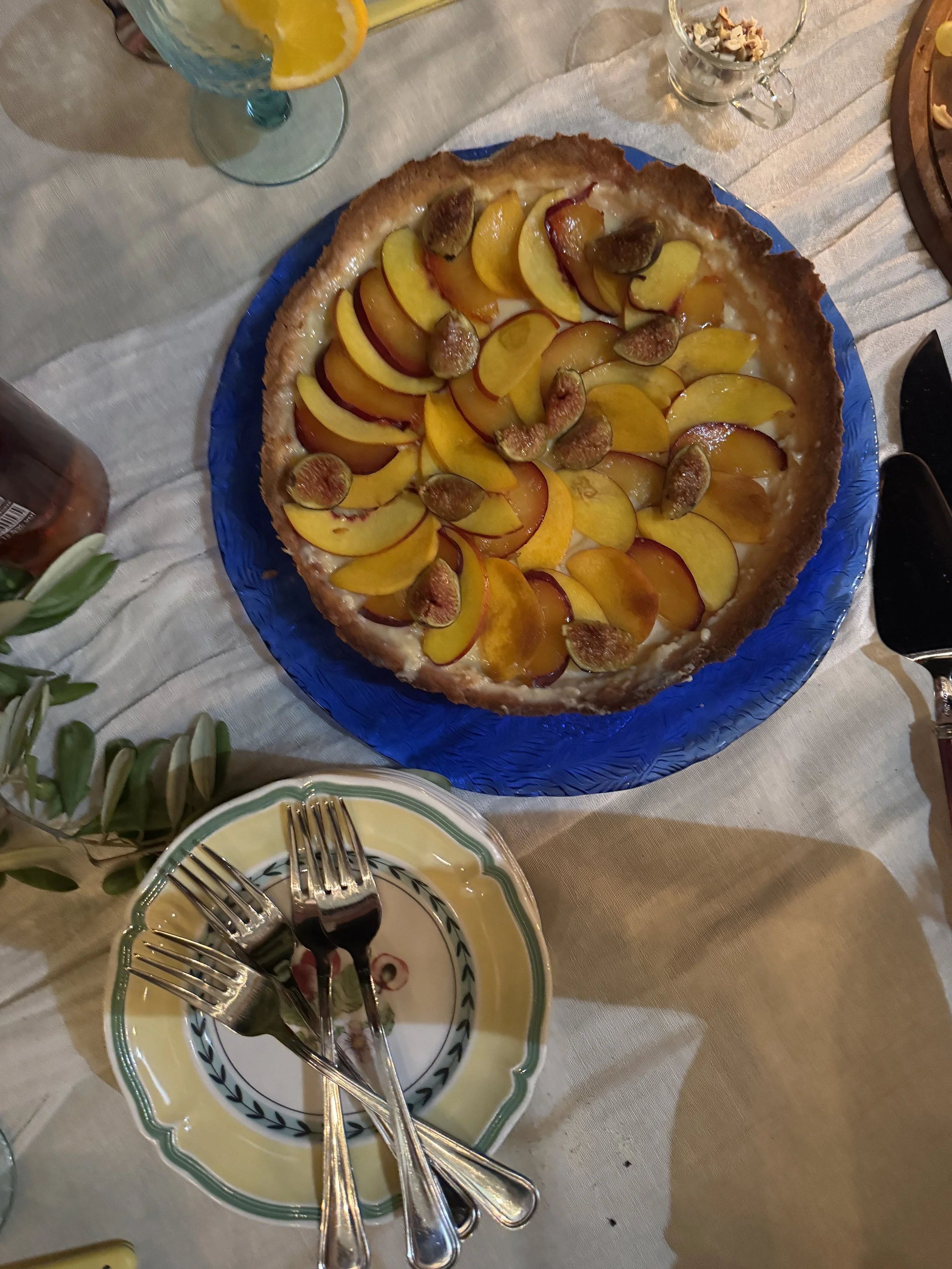 Peach tart on a blue plate at a dining table with slices of peach and figs on top, surrounded by a plate with several forks, a glass of lemon water, and other tableware.