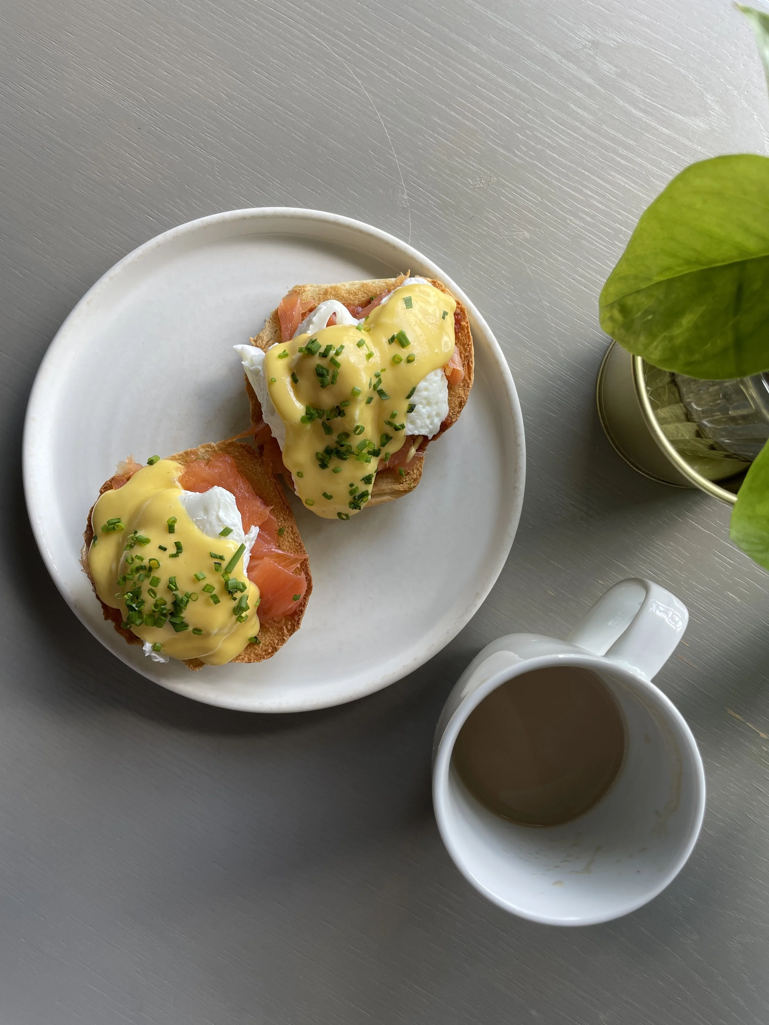 Two eggs Benedict with smoked salmon on toast, topped with hollandaise sauce and chopped chives on a white plate, with a cup of coffee and a potted plant nearby.