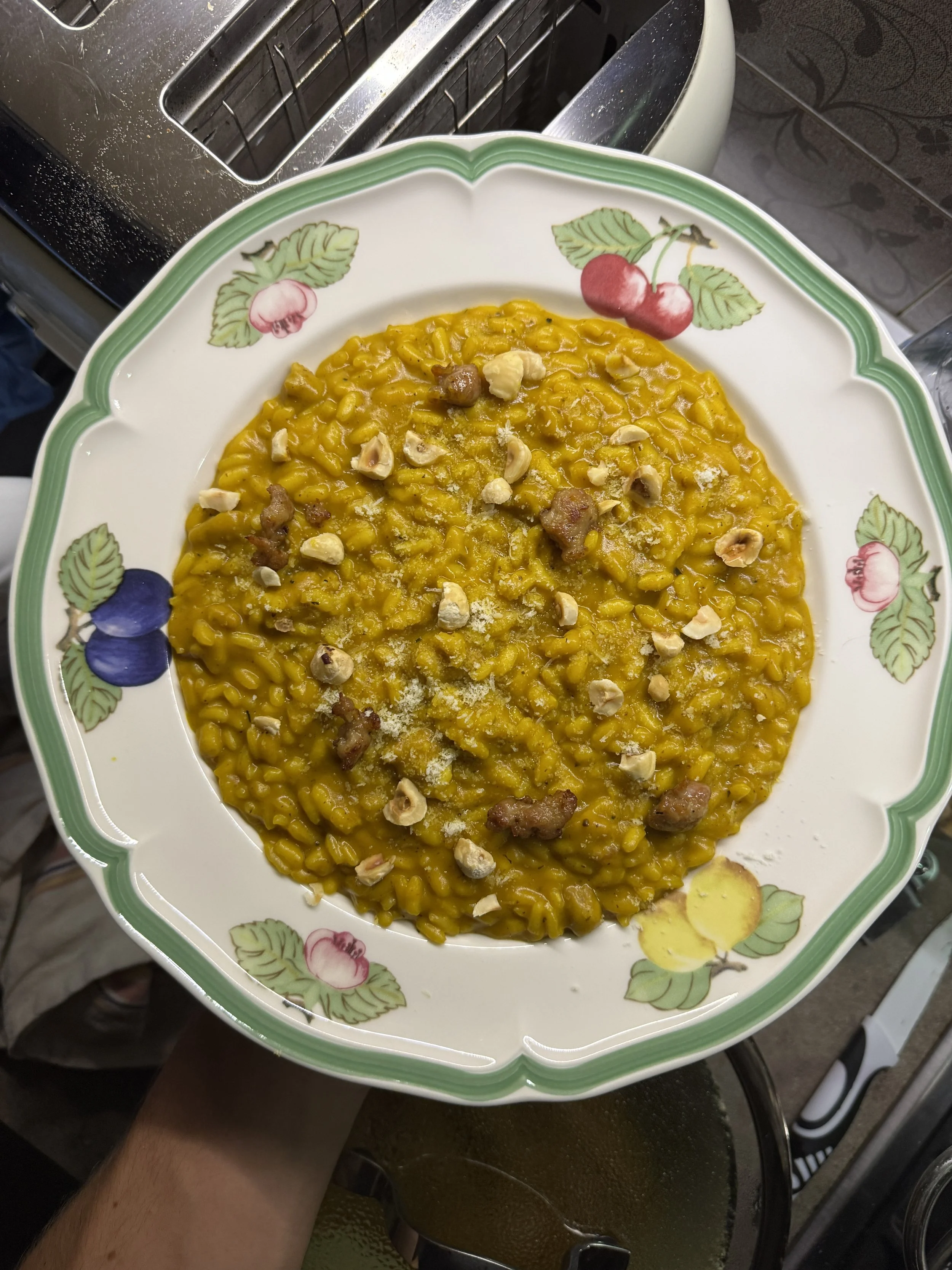 A decorative plate of yellow risotto garnished with chopped nuts. The plate has a floral pattern with cherries, blueberries, and lemons around the border. The plate is held over a kitchen counter with a toaster in the background.
